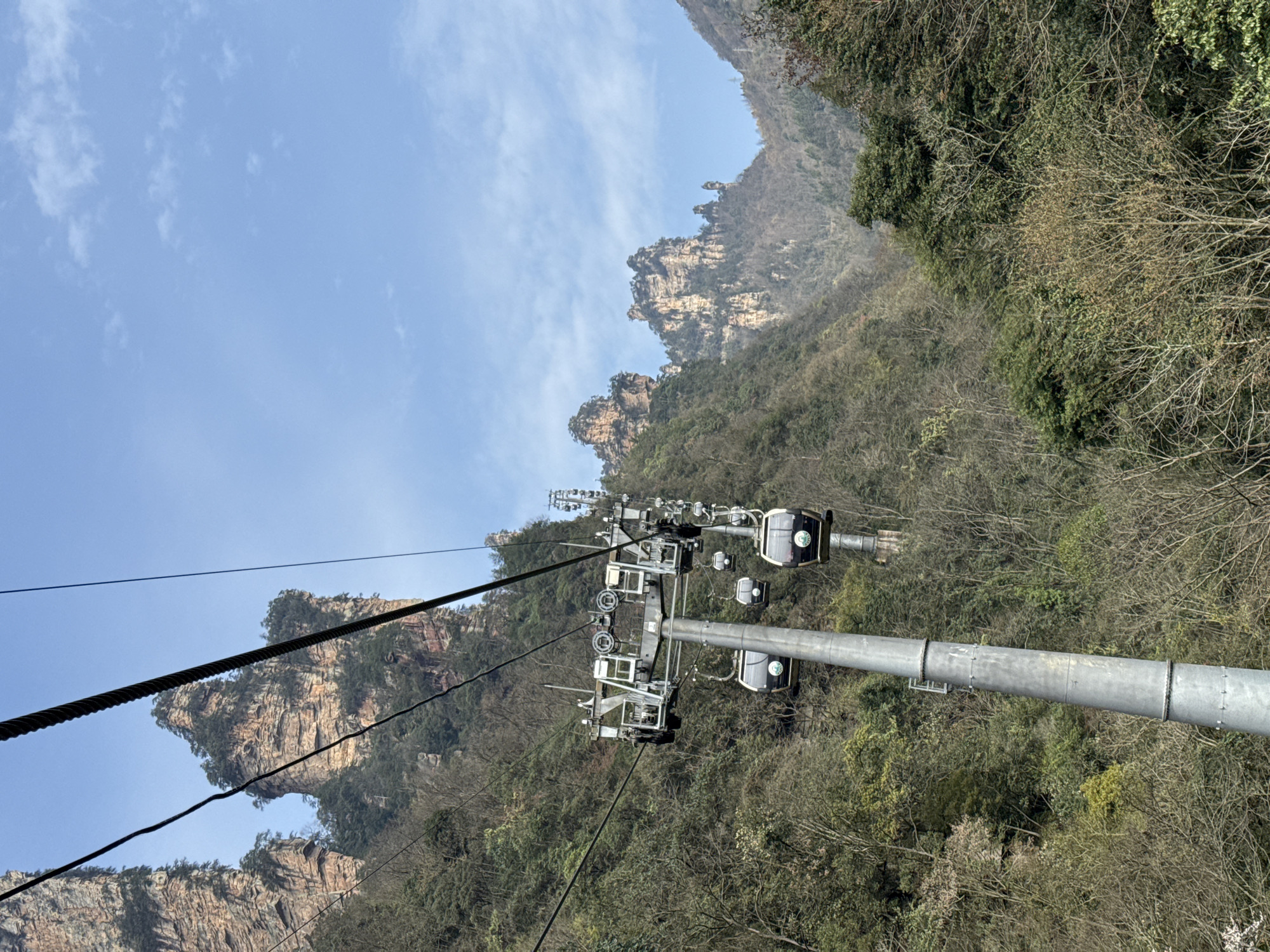 This image depicts a section of a gondola lift system within the Wulingyuan scenic area, China. A prominent grey metallic pylon, appearing to be steel, stands vertically in the left-center foreground, serving as a support tower for multiple parallel steel cables. Three dark-grey gondola cabins, featuring lighter tops and a circular green emblem on their sides, are suspended from these cables near the pylon. Additional similar gondola cabins are visible in the distance along the cable system, extending over the mountainous terrain. The surrounding environment consists of steep mountain slopes densely covered with a mix of deciduous trees, many appearing bare or with sparse foliage, and interspersed evergreen vegetation. Numerous towering, sheer-sided sandstone pillars, characteristic of the Wulingyuan karst topography, rise majestically in the midground and background. These pillars exhibit stratified rock formations and sporadic patches of greenery. The sky above is clear and light blue, with minimal, dispersed white clouds. No individuals are visible in the scene.
