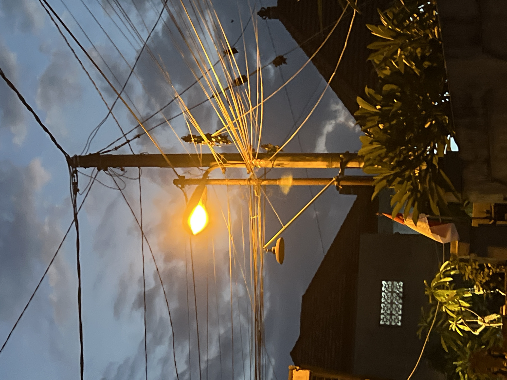 The image captures a street scene at dusk in Denpasar, Indonesia. A tall, wooden utility pole dominates the center, heavily laden with a dense network of electrical wires that stretch across the frame. One of the wires is illuminated by a streetlamp, casting a bright yellow glow on its surface and on the surrounding cables. Below the main streetlamp, a second, smaller lamp hangs, also illuminated, and angled downwards.
In the lower left of the image, the dark, sloped roof of a building is visible, with a single window featuring a patterned grate. To the right of this building, a flag with red and white bands, likely the Indonesian flag, is partially visible, attached to a pole. In the foreground, lush green foliage obscures the lower portion of the scene, illuminated by the ambient light. The sky above is a deep blue, tinged with grey clouds, indicating twilight or a cloudy evening. The overall impression is a somewhat cluttered but typical urban streetscape as night begins to fall.