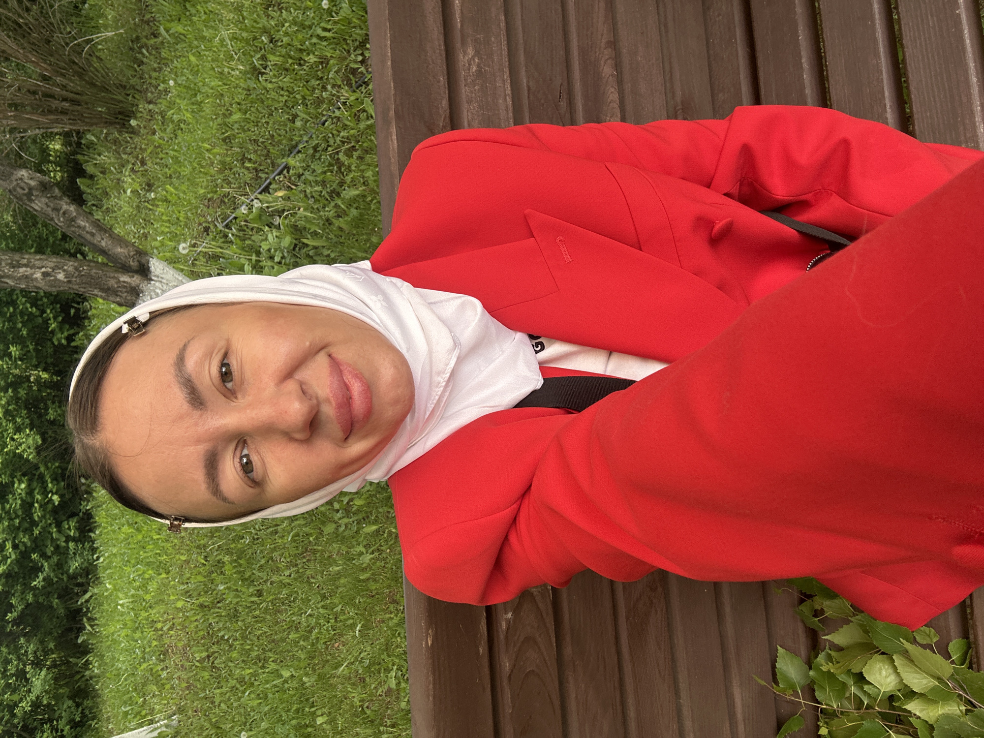 A woman wearing a white headscarf, red blazer, and pants is sitting on a wooden bench in a grassy area. The location is likely outdoors, possibly in a park or garden in the city of Votkinsk, Russia, based on the request. The woman is looking at the camera and smiling slightly.