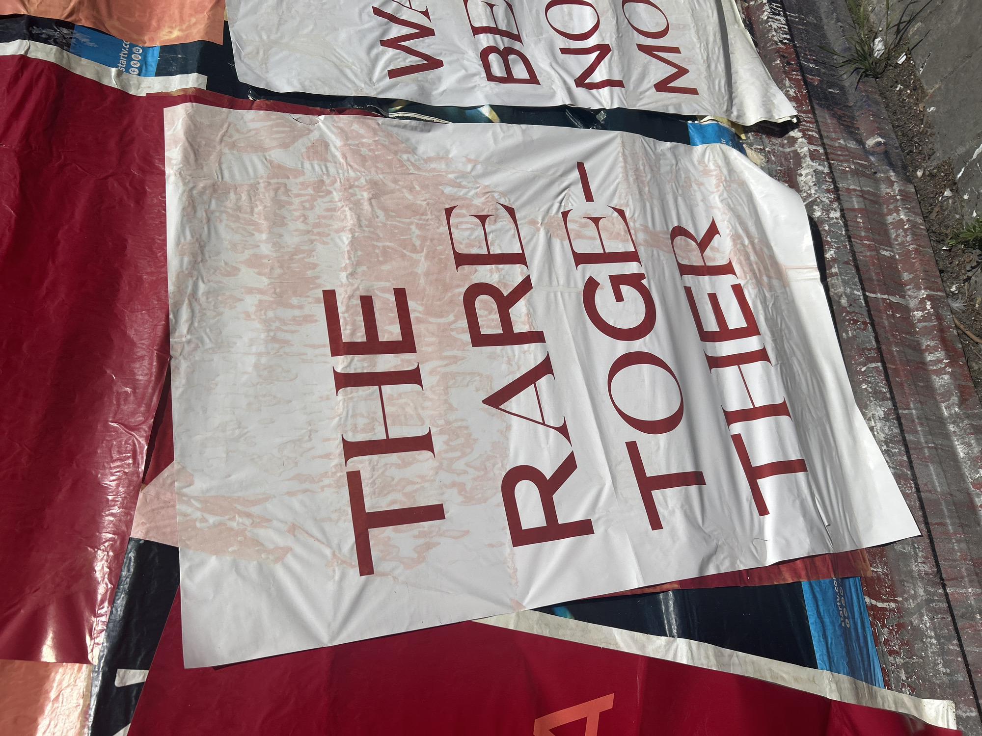 The image shows discarded advertising posters lying on the ground, likely in an urban area. The poster in the foreground reads "THE RARE TOGETHER" in large, red serif font. Another poster partially visible above reads "WE BELONG MORE." The posters are torn and appear worn. The ground is partially paved and shows signs of wear and dirt. Beyoğlu, Turkey, is not explicitly apparent, but a crowded, urban setting is indicated.