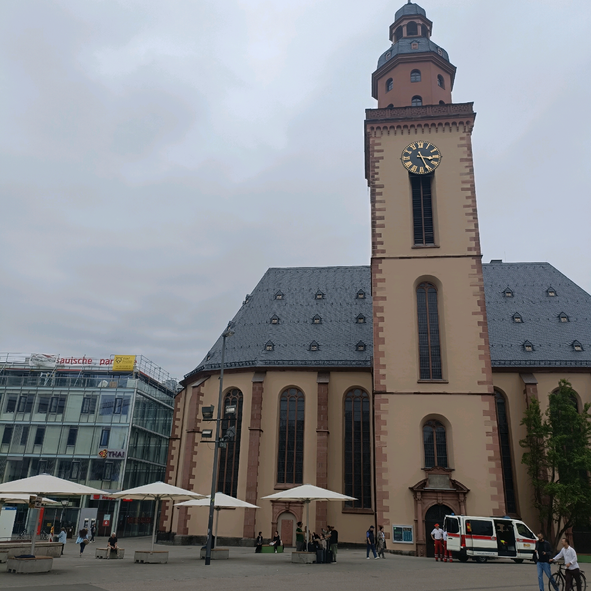 This image captures a scene in the city of Frankfurt, Germany, focusing on the historic Katharinenkirche (St. Catherine's Church) and its surroundings. The Katharinenkirche, a prominent landmark, stands tall with its beige facade, reddish-brown stone accents, and a dark grey slate roof. Its steeple is topped with a grey spire, and a large clock face with Roman numerals is visible on its tower. Adjacent to the church, a modern glass-fronted building with a multi-level structure is under construction or renovation, indicated by scaffolding and banners. The area in front of the church and the modern building appears to be a public square, paved with grey stone. Several white umbrellas are set up, suggesting an outdoor seating or market area. People are scattered throughout the square, some walking, some sitting on benches. An ambulance is parked near the entrance of the church, with its doors open, and two individuals in what appear to be medical uniforms are standing nearby. The sky is overcast, contributing to a diffused lighting condition across the scene. The overall impression is a blend of historic architecture and contemporary urban development in a bustling city center.