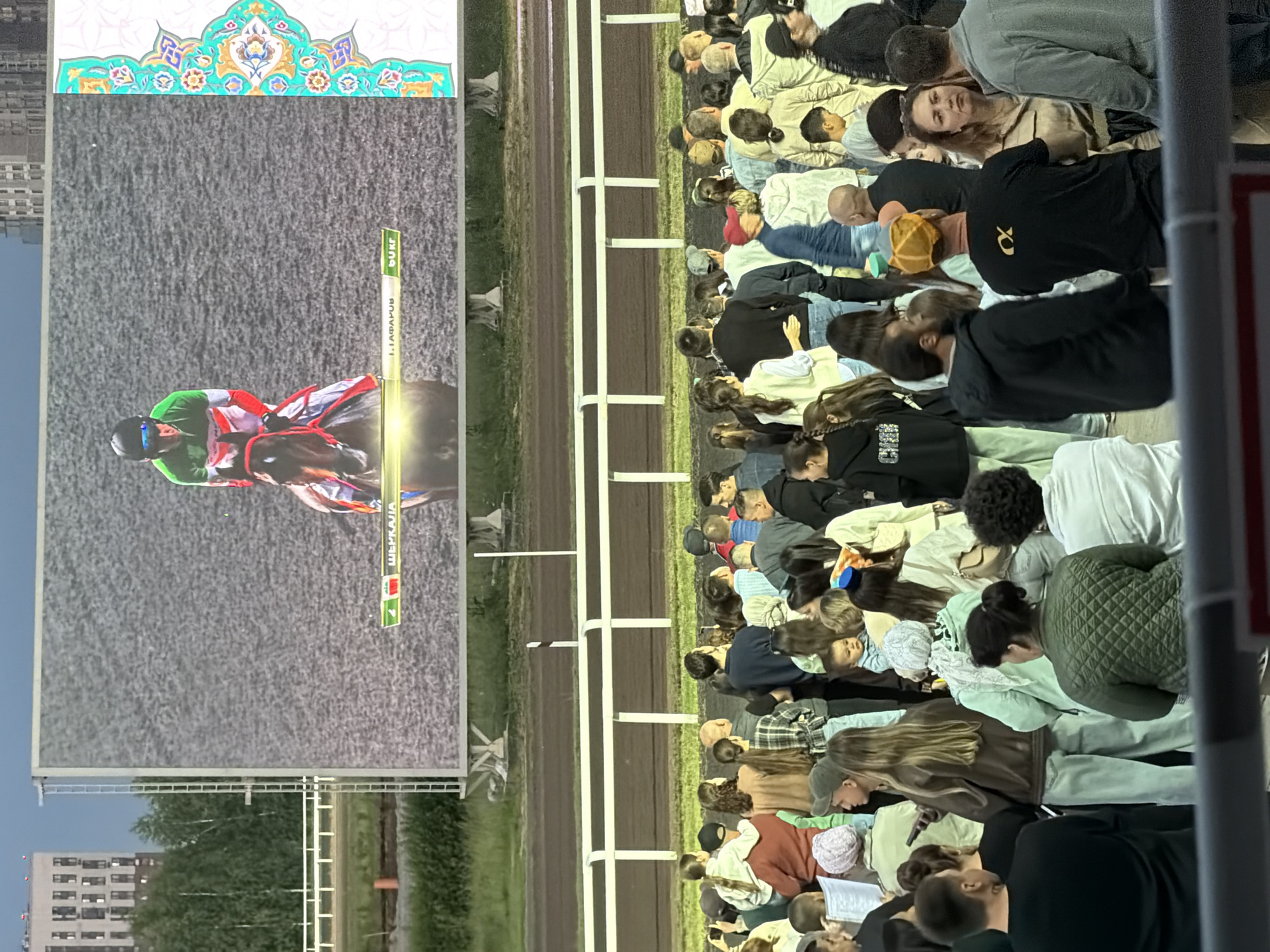 A large outdoor screen displays a horse race with a jockey in a green, white, and red outfit. Below the screen, a crowd of people are gathered, facing the screen. The environment appears to be a race track, with a fence and track visible in the background. The sky is overcast and it seems to be evening or late afternoon.