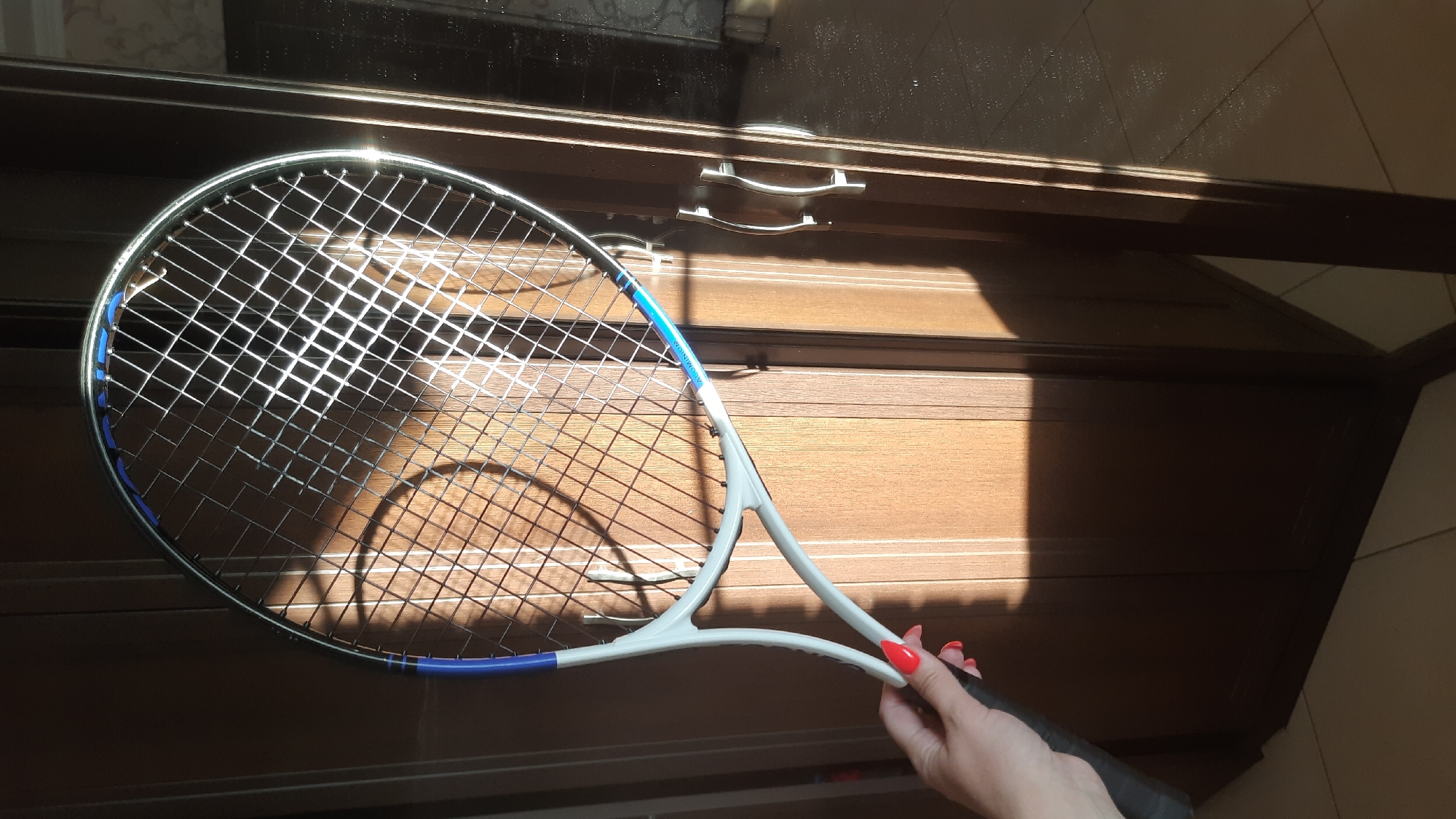 A person is holding a tennis racket in front of a wooden wardrobe. The racket has a white frame with blue accents and silver strings. The wardrobe has three doors and is made of dark wood with vertical paneling. Sunlight is casting shadows across the scene, creating bright highlights on the racket strings and the wardrobe doors. The floor appears to be tiled. The image was taken in Desnogorsk, Russia.
