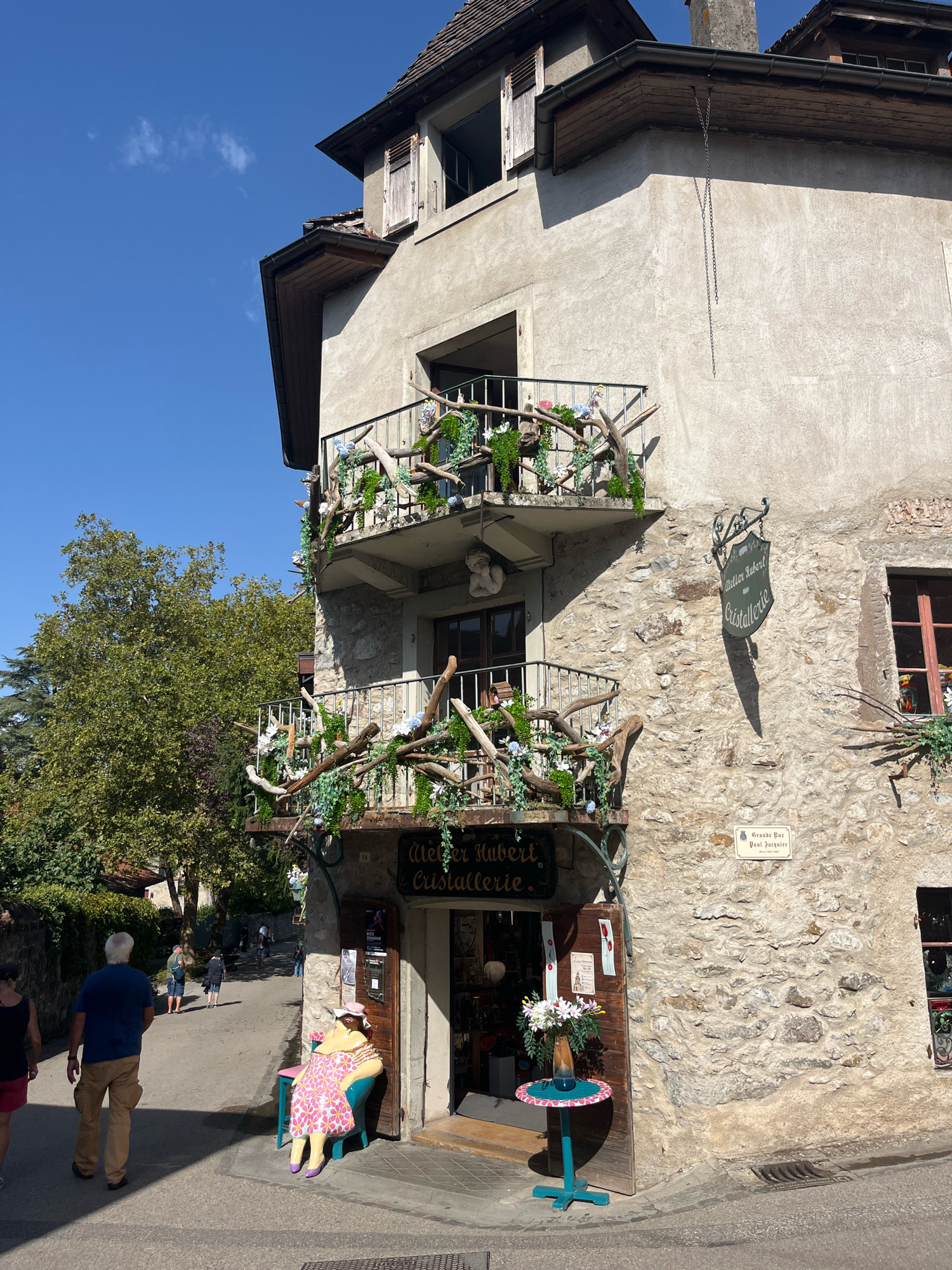 The image depicts a charming corner building in the medieval town of Yvoire, France, bathed in bright sunlight. The building features stone walls and balconies adorned with rustic wooden branches and artificial greenery and flowers. A prominent sign hanging from the wall reads "Atelier Hubert Cristallerie," indicating a crystal workshop. Below the balconies, an open doorway reveals the interior of the shop, with items displayed inside. To the left of the entrance, a large, colorful mannequin of a woman sits on a turquoise chair. A small, teal table with a floral arrangement stands in front of the shop. In the background, a cobblestone street curves upwards, with several individuals walking and a lush green tree. The sky is clear and blue.