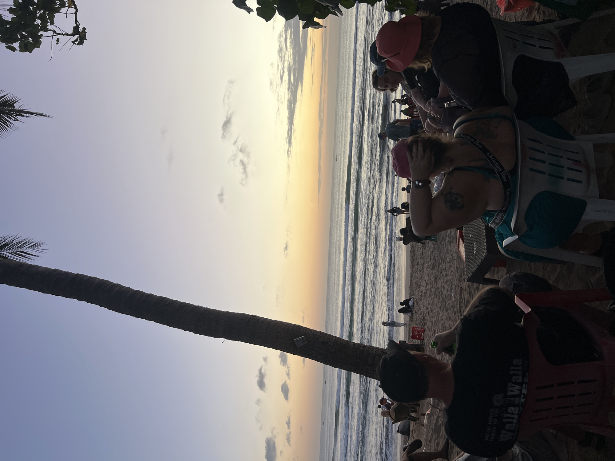 The image depicts a beach scene in Kuta, Indonesia, during sunset. A tall, dark palm tree dominates the left side of the frame, leaning over a sandy beach where several people are seated in plastic chairs. In the foreground, a man wearing a black t-shirt and a baseball cap is holding a green bottle and facing away from the camera, seated in a red plastic chair. To his right, two women are seated in white plastic chairs, facing the ocean. The woman on the left wears a pink hat and a teal top, with her hands behind her head. The woman on the right wears a red hat and a dark top.
The beach is populated with other individuals scattered around, some standing and some seated. The ocean in the background displays gentle waves, and a few small boats are visible on the horizon. The sky is a gradient of pale blue at the top, transitioning to soft oranges and yellows near the horizon, indicating the setting sun. The overall atmosphere suggests a relaxed, casual evening gathering on the beach.