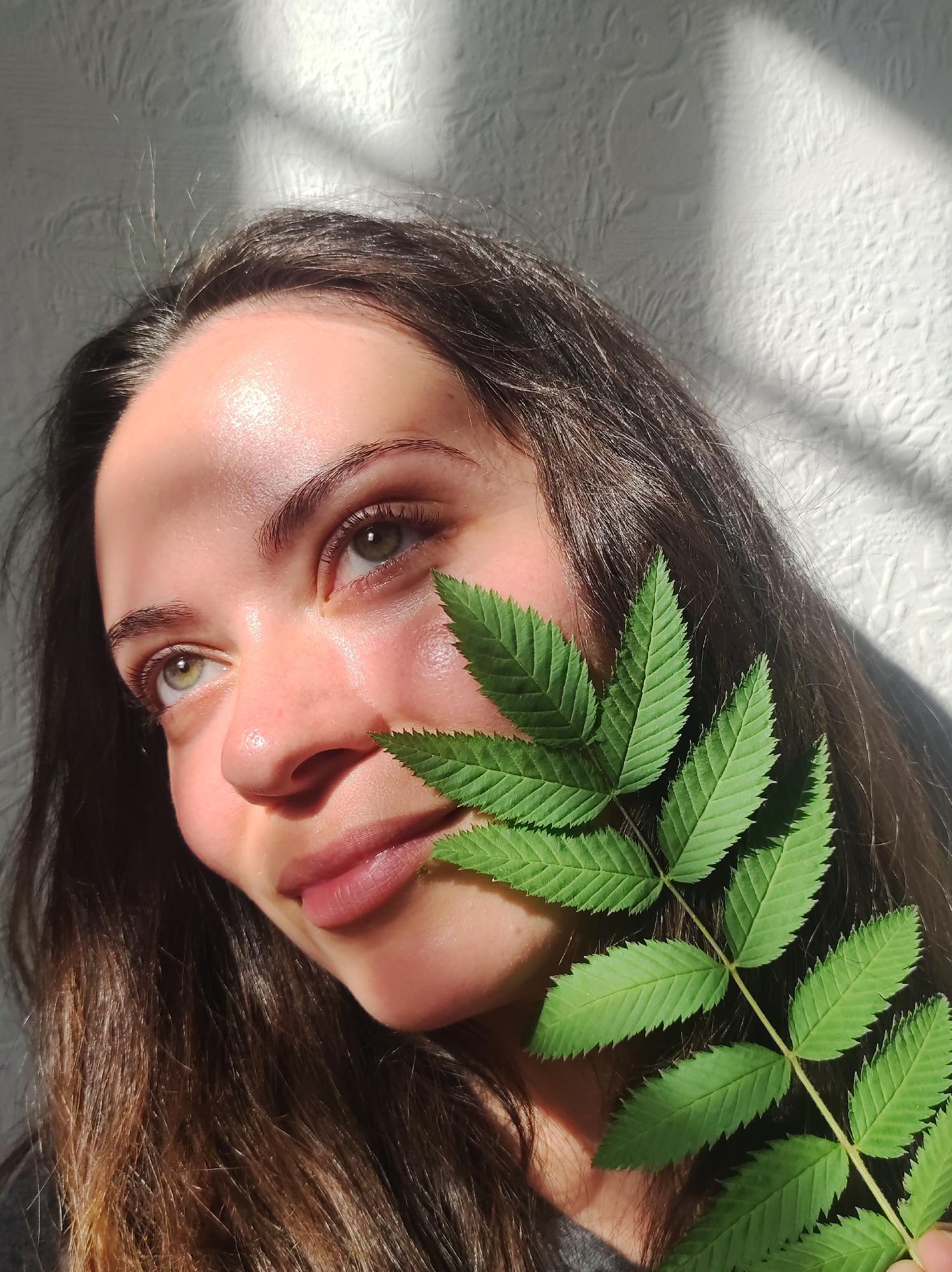 A close-up portrait features a young woman with long, brown hair. A bright green leaf with multiple leaflets covers part of her face, obscuring her cheek and part of her nose. Her skin is fair and illuminated by strong, natural light, creating highlights on her forehead and cheek. She has light eyes and wears subtle lip color. The background is a textured white surface, likely a wall, with a distinct shadow pattern cast upon it.
It is not possible to determine the exact location of the photo with the information. It is not possible to know if it was taken in the city of Chaikovsky, Russia.