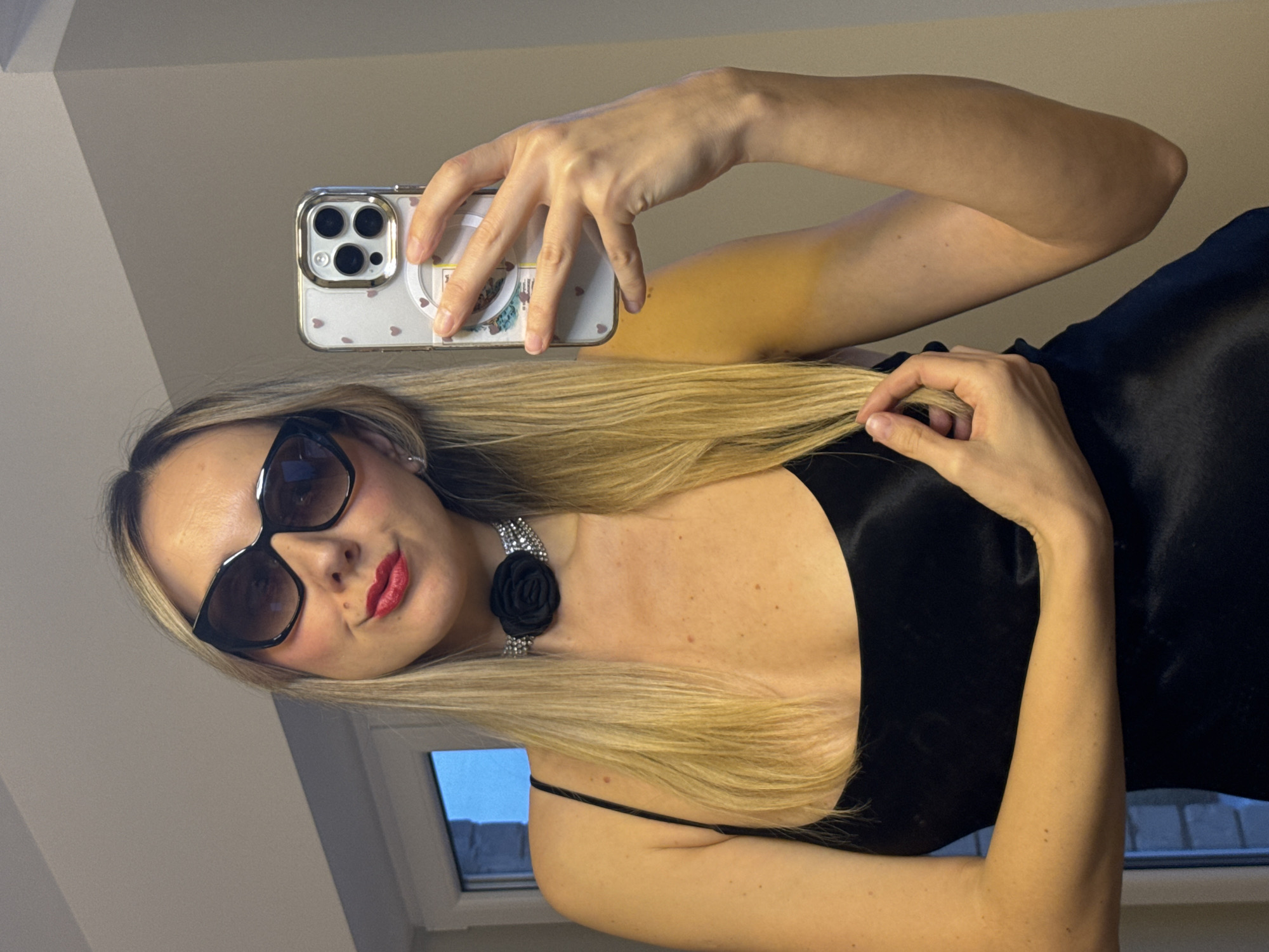 A woman is taking a selfie. She is wearing sunglasses, red lipstick, and a black dress with a black rose choker. She is holding her phone in her left hand, and her right hand is touching her hair. The background is a plain wall and part of a window with the outside visible through it. It's impossible to determine if this photo was taken in Carnikava, Latvia from the visual information provided.
