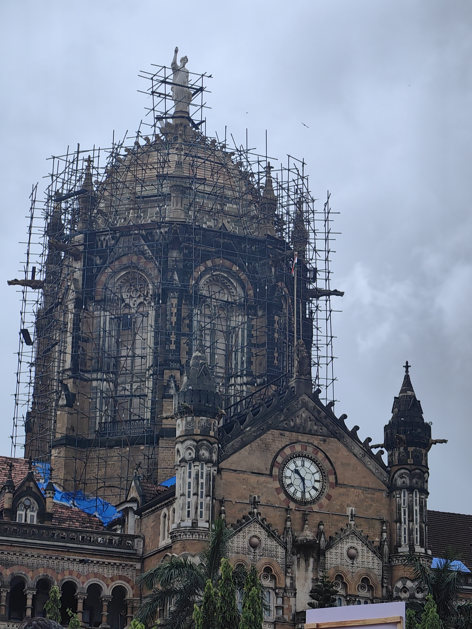 The image depicts a close-up, low-angle view of the Chhatrapati Shivaji Maharaj Terminus (CSMT) in Mumbai, India. The historic railway station, a UNESCO World Heritage Site, is undergoing extensive scaffolding work, encasing its iconic dome and upper structures. The dome itself is covered in scaffolding, with a statue of a figure, possibly representing progress or a historical emblem, visible at its apex. Decorative elements, architectural details, and windows are partially obscured by the scaffolding. To the right, a large clock face with Roman numerals is prominently displayed on the building's facade, showing the time as approximately 10:09. Several spires and decorative gargoyle-like figures are visible jutting out from the stone architecture. Blue tarpaulin covers some sections of the roof. In the foreground, the tops of trees, likely palm trees, are visible. The sky is overcast and appears grayish, suggesting cloudy weather. No people are clearly visible in the frame, though a glimpse of what might be a person's head or shoulder is at the very bottom of the image. The overall scene conveys a sense of ongoing restoration and preservation of a significant historical landmark.