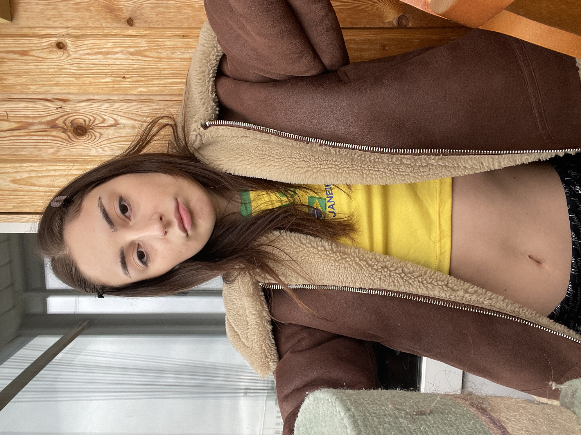 A young woman with long, dark brown hair and light makeup is captured in a close-up, upward-angled shot. Her head is tilted to her right, and she looks directly at the camera with a neutral expression. She is wearing a brown faux shearling jacket with a cream-colored fleece lining visible along the collar and zipper. Beneath the open jacket, she wears a bright yellow crop top, exposing her midriff where a small navel piercing scar is visible. The yellow top features a partially visible graphic that includes green elements and the word "JANEIR", likely referring to Rio de Janeiro, suggesting a Brazilian theme. Below the crop top, she wears black patterned shorts or pants. A small, dark hair clip is visible in her hair above her left temple.
The setting appears to be indoors, against a backdrop of light wooden wall paneling with prominent grain and knots. To her left, there is a window or glass door with horizontal blinds or slats, allowing soft light into the space. In the foreground, part of a light-colored, possibly knitted or woven, blanket or fabric with faint striped patterns is visible. The overall scene suggests a casual indoor environment, possibly a home, in Moscow, Russia. The lighting is natural, likely from the window, indicating daytime.
