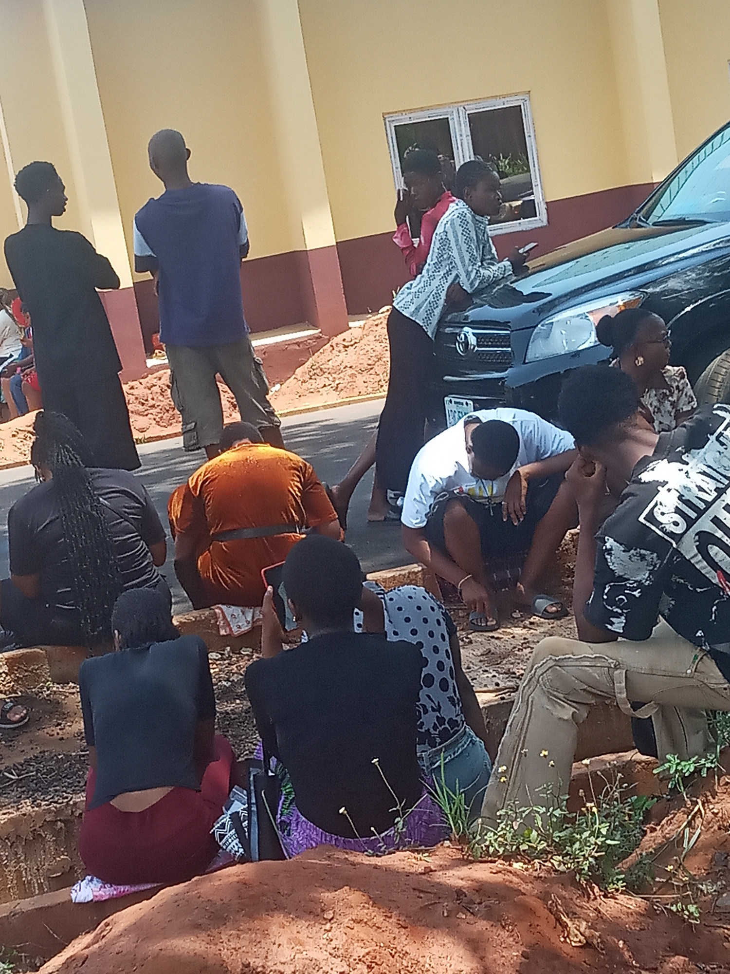 In Ukwu-Abwa, Nigeria, a group of people are gathered on a roadside area with exposed red soil and vegetation. Some individuals are seated on what appears to be a low earth embankment or curb, while others stand nearby. The group is situated next to a black SUV parked along the road, with a light-colored building visible in the background. Some people are looking down at something on the ground. At least one person is holding a phone, seemingly taking a photo. People wear a variety of casual clothes.