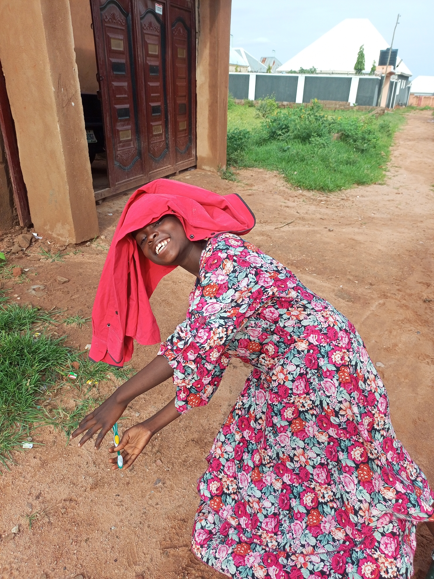 In Tempure, Nigeria, a young Black girl with dark skin and short black hair is captured in a medium shot. She is wearing a vibrant floral dress and a red garment draped over her head, resembling a makeshift headscarf. The girl is leaning forward, her body angled towards the left of the frame, and she is smiling broadly with her mouth open. Her left arm is extended, and her hand is reaching towards the ground. In her right hand, she holds a green and yellow toothbrush. The background consists of a dirt path, sparse green grass, and a portion of a brown building with a large, dark wooden gate. The lighting suggests an overcast day.