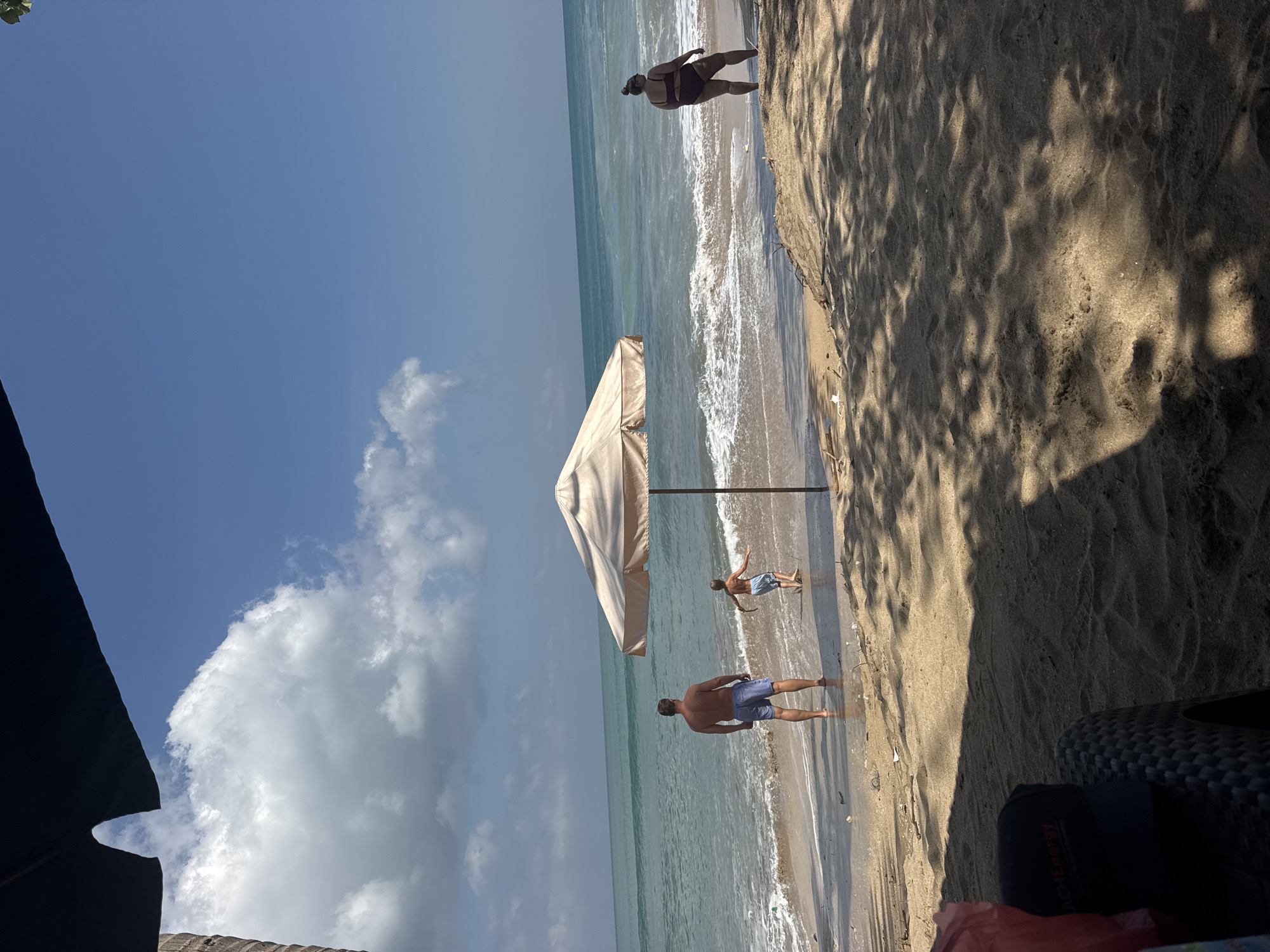 A wide shot captures a sun-drenched beach scene in Kuta, Indonesia. The foreground is dominated by sandy ground with dappled sunlight and shadows from overhead foliage. A dark-colored beach chair with a woven texture is visible on the left, with a black baseball cap resting on it. A red bag is also present.
The midground features the shoreline with gentle waves lapping the sand. A large, cream-colored beach umbrella stands near the water. Three individuals are present on the beach. A man wearing blue swim trunks stands facing the ocean, his back to the camera. A young boy in blue swim trunks is running in the shallow water. On the right, a woman in a dark-colored swimsuit stands facing away from the camera, looking towards the ocean.
The background consists of the turquoise ocean meeting a clear blue sky with scattered white clouds. The overall impression is a peaceful day at the beach.