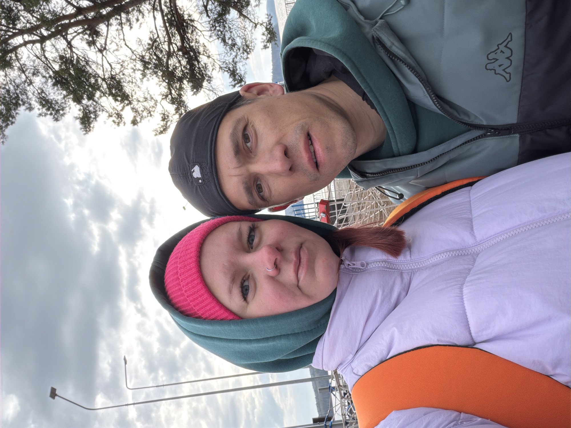 Two people pose for a selfie. A man wears a dark baseball cap and a Kappa brand jacket. A woman wears a pink knit hat under a hooded jacket. The backdrop includes trees, a cloudy sky, and an apparent body of water. The location is likely Chaikovsky, Russia. Streetlights are visible in the background.