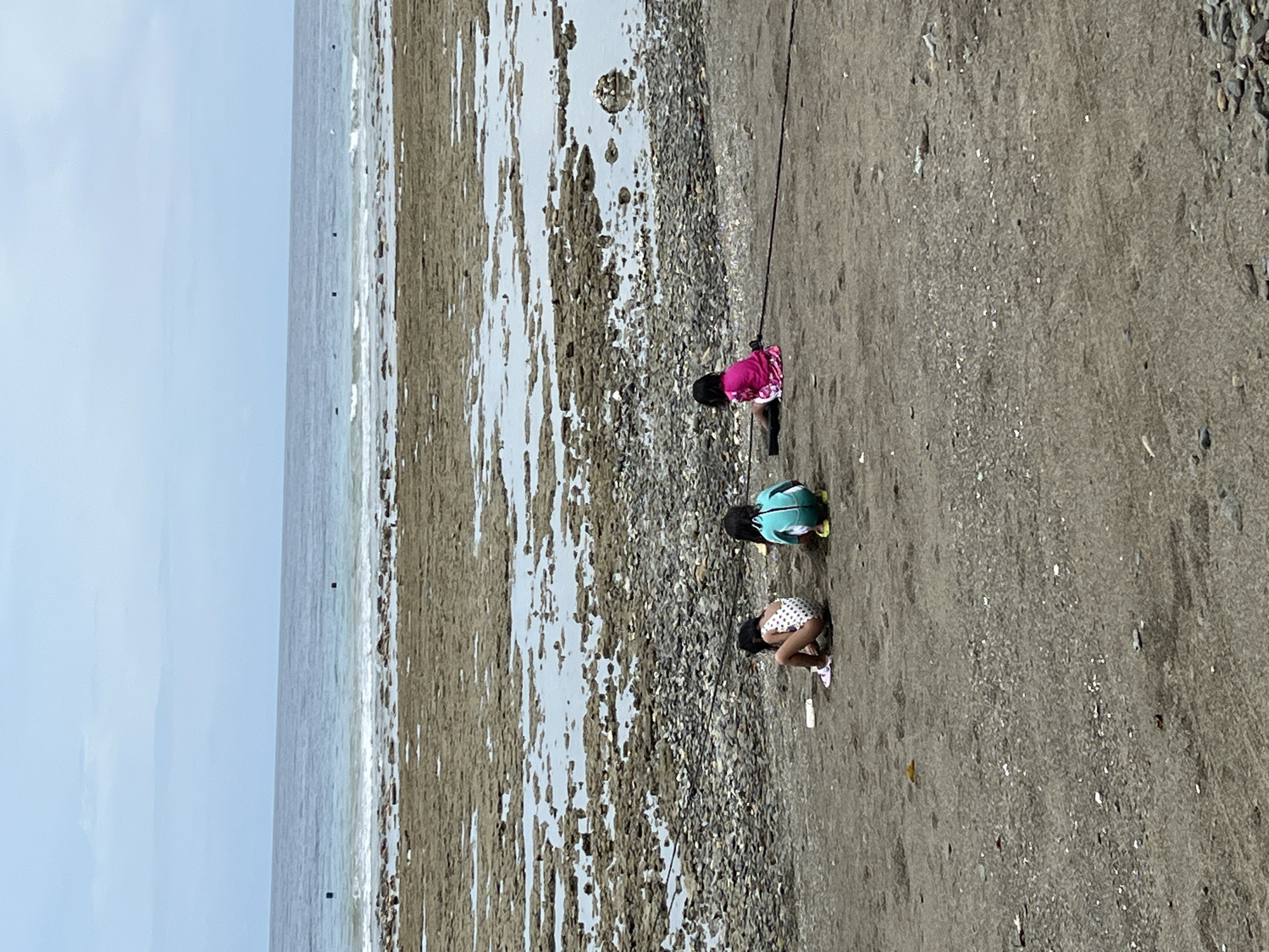 Three children are crouching on a beach. The child farthest from the camera wears a pink shirt and dark bottoms. The child in the middle wears a teal shirt and light bottoms. The child closest to the camera wears a white dress with black polka dots. The beach extends from the bottom right to the middle left of the frame, where it meets the ocean. The sky is visible in the top left corner of the frame. The location is in Banaybanay, Philippines.