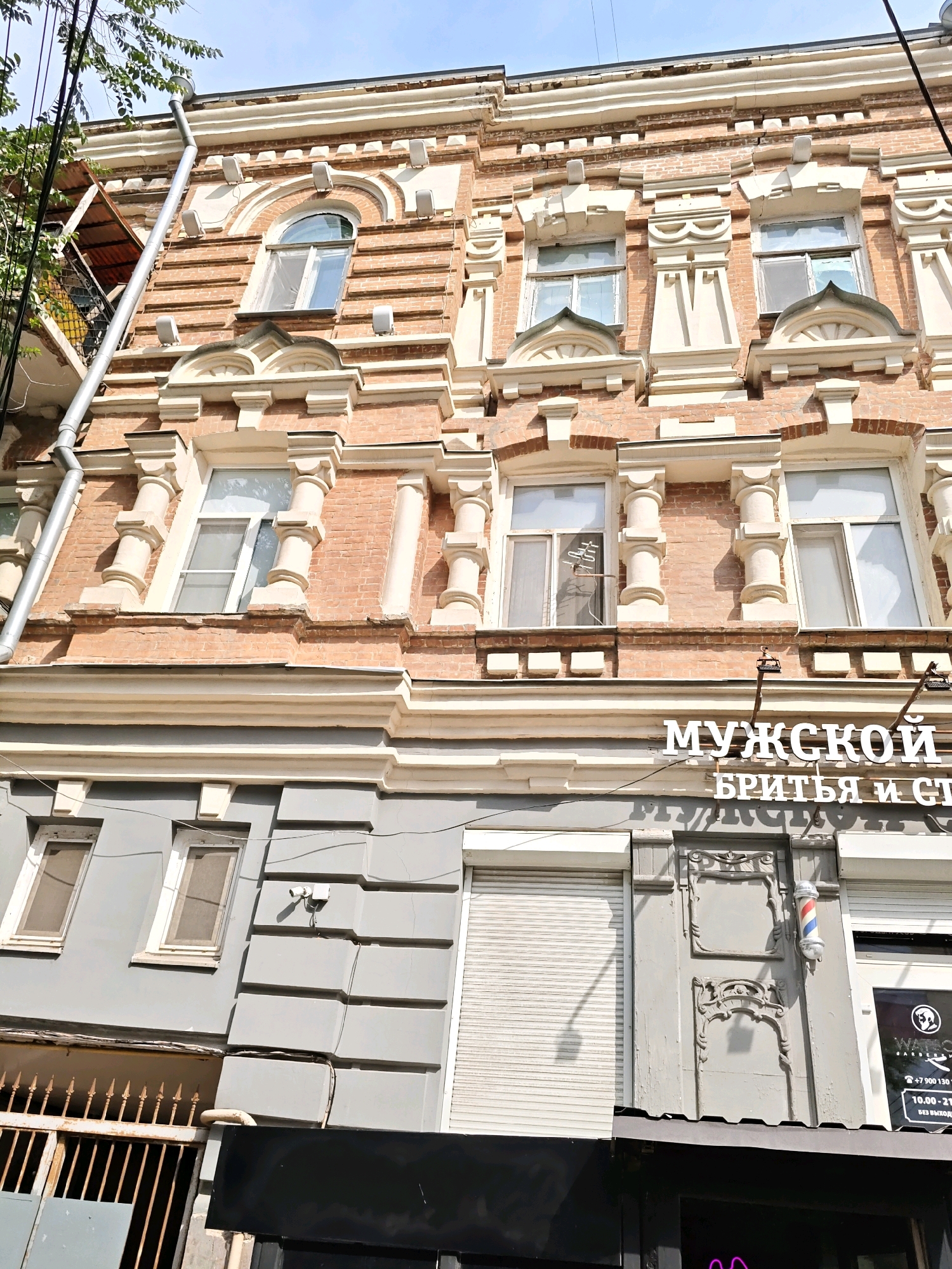 The image shows the facade of a building in Rostov-on-Don, Russia. The building has an ornate, multi-story design with brick and plaster elements. On the lower part of the building, there is signage in Russian that reads "МУЖСКОЙ БРИТЬЯ И СТ" which translates to "Men's Shaving and C". Below this sign, there is a barber pole, indicating that this is likely a barbershop. The building appears to be a mix of old and new, with modern security cameras and rolling shutters visible alongside the historical architectural details. The sky is blue and clear, suggesting it is daytime. No people are clearly visible in the image.
