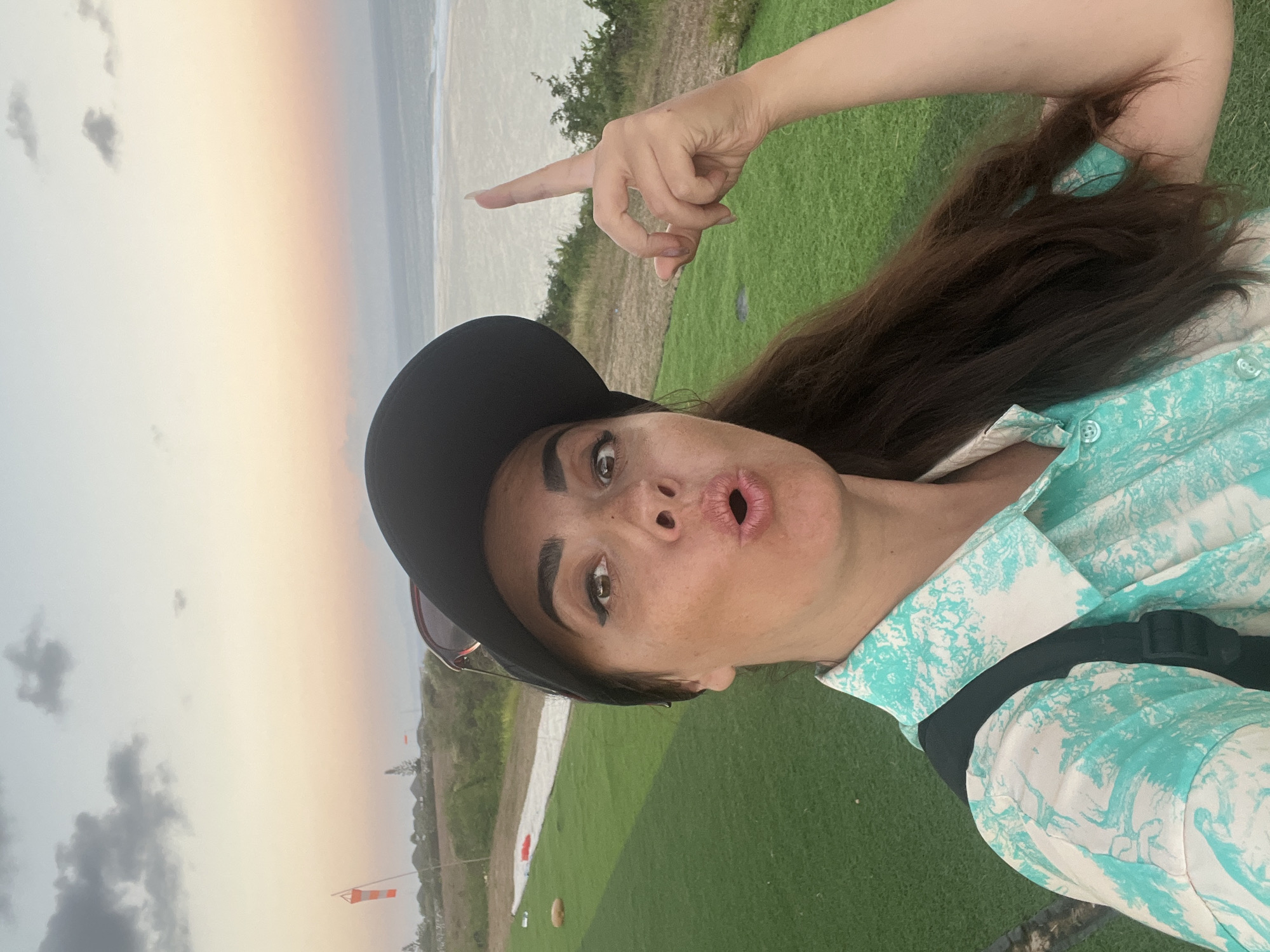 A selfie of a woman with long brown hair and brown eyes, wearing a black baseball cap, sunglasses, and a teal and white patterned button-up shirt. She has her mouth open in a playful expression and is pointing upwards with her right index finger. The background features a green grassy area, a runway or landing strip, a windsock, and the ocean with waves under a hazy, pastel-colored sky at dusk. The location is identified as Kuta Selatan, Indonesia.