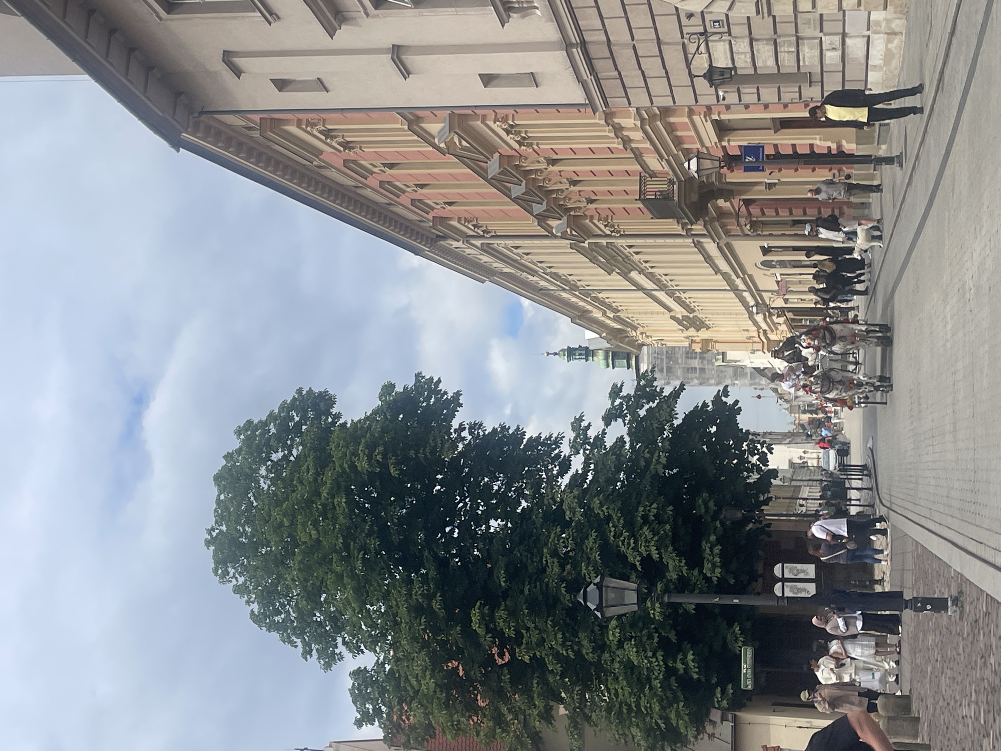 The image depicts a street scene in Krakow, Poland, likely in the Old Town or Kazimierz district, characterized by historic architecture and a cloudy sky. On the left, a large, leafy green tree dominates the foreground, partially obscuring a lamppost. A group of people, some casually dressed, are gathered near a building entrance. Further down the street, a horse-drawn carriage is visible, with passengers seated and a driver. The street itself is paved with cobblestones, and lined with buildings featuring classical architectural elements, such as decorative facades, windows, and balconies. Several other people are seen walking along the sidewalk. The overall atmosphere suggests a day of tourism or leisurely activity.