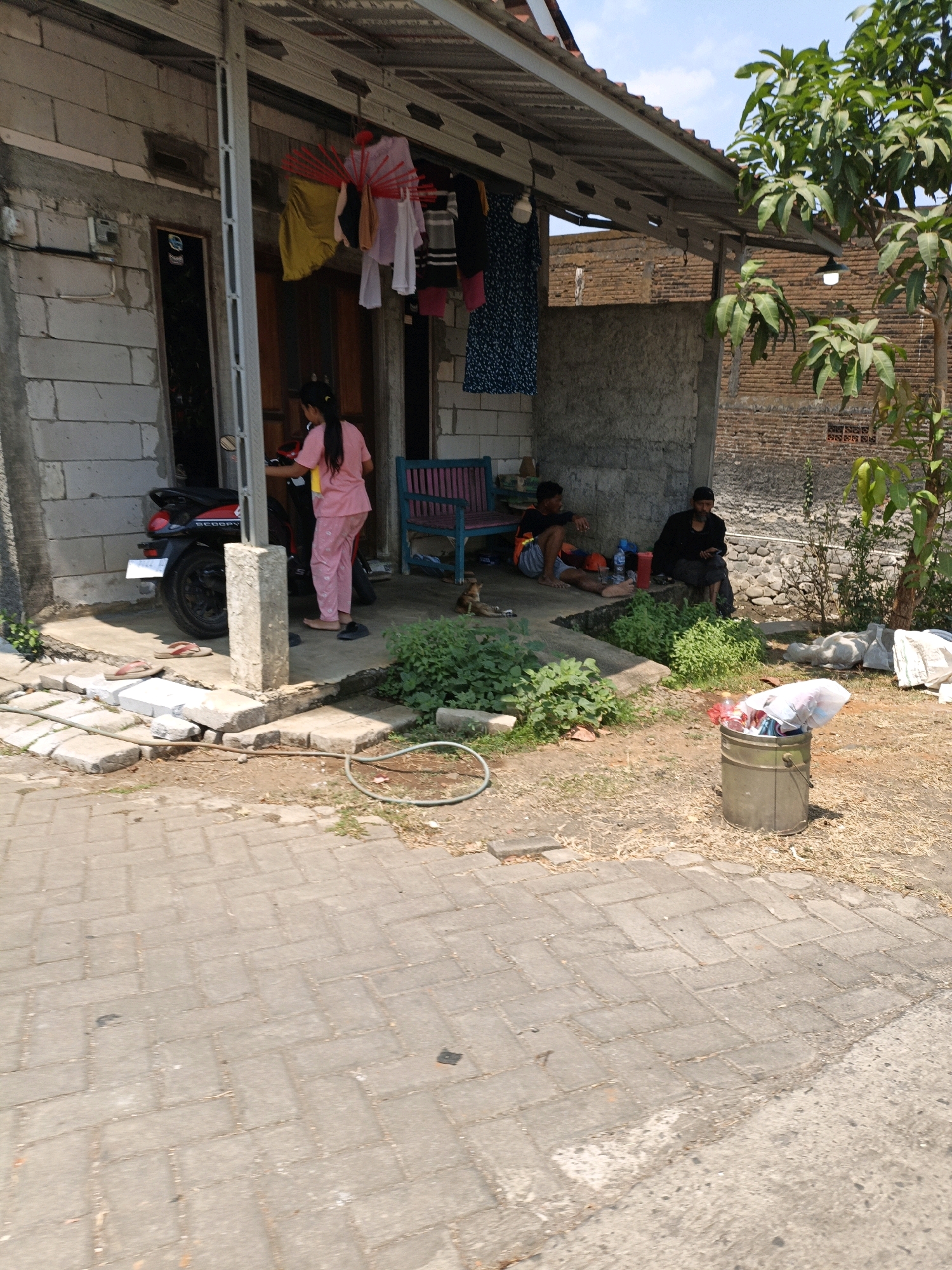 This image depicts a residential scene in Jepara, Indonesia, likely a modest home with an open-air front. In the foreground, a paved walkway leads towards the house. A woman in pink pajamas is standing near a red and black motorcycle parked on the concrete area in front of the house. Clothes are hung out to dry on a line extending from the porch roof. Further back, two men are sitting on the ground under the porch. One man, wearing a black shirt and headscarf, is seated against a wall. Next to him, a younger man in a t-shirt and shorts is also sitting on the ground. Lush green plants grow in front of the porch, and a metal trash can filled with various items is visible in the middle ground. The house itself appears to be constructed with cinder blocks and has a corrugated metal roof. The background reveals a brick wall and trees, suggesting a semi-urban or rural environment. The overall impression is one of everyday life in a tropical climate.