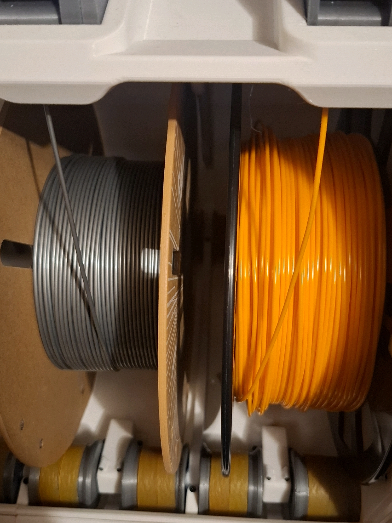 The image shows two spools of filament, one gray and one orange, inside a 3D printer or similar device. The spools are stacked vertically, with the gray spool on the left and the orange spool on the right. Both spools are full, with the filament tightly wound. A thin gray filament from the gray spool is visible extending upwards. A thin orange filament from the orange spool is also visible extending upwards. The spools are held in place by a mechanism that includes yellow and gray cylindrical components at the bottom of the image. The background is a white housing with some gray elements visible at the top. There is no indication of the location in the city of Tyumen, Russia, within the image itself.