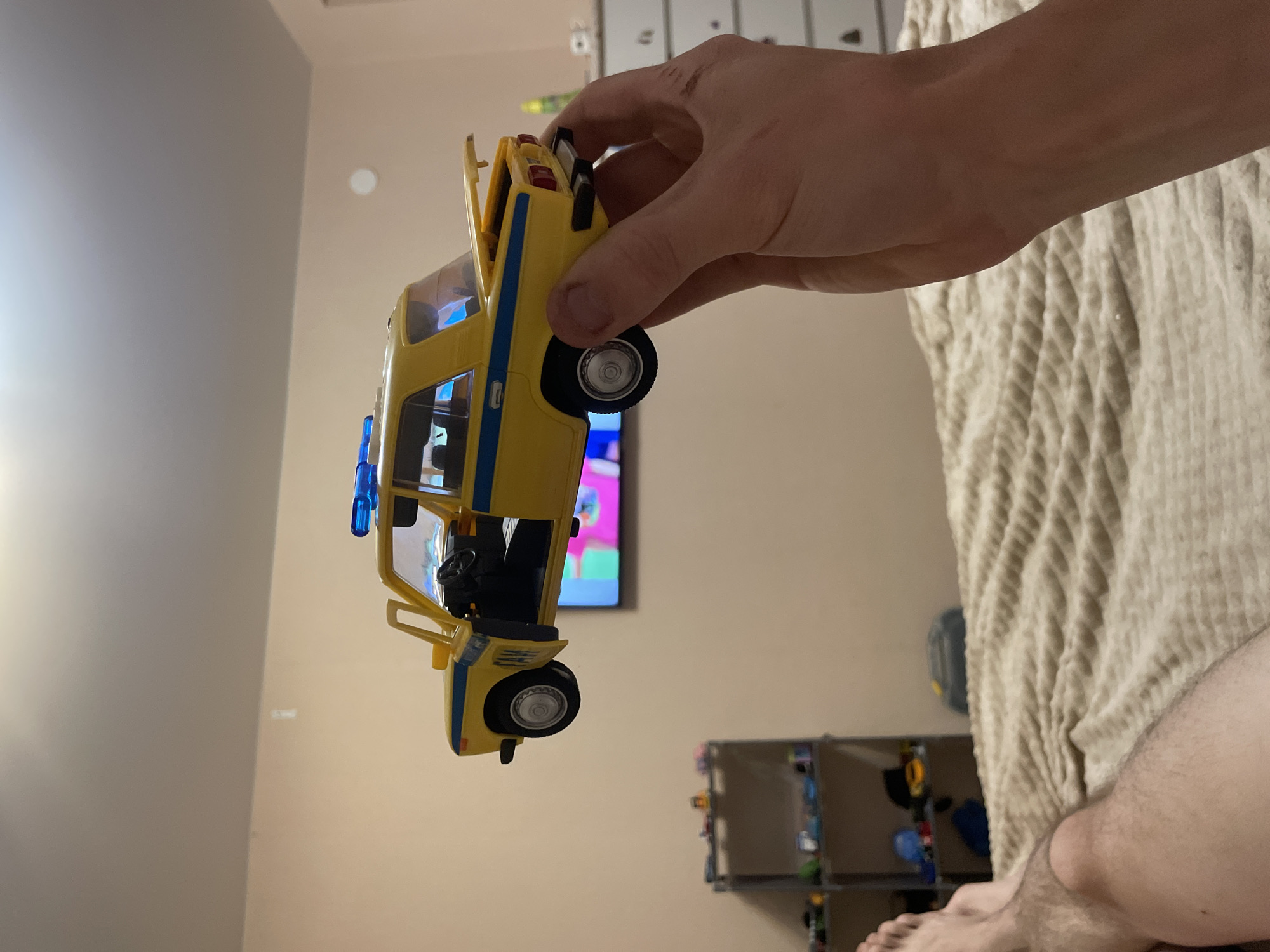 A hand is holding a yellow toy van with blue stripes. The van is depicted from the side, with its doors open, revealing a black interior and a steering wheel. The van has blue flashing lights on its roof. The background consists of a beige wall and a patterned beige blanket, suggesting an indoor setting, possibly a bedroom. A television screen displaying a colorful image is visible in the background. In the lower right corner, a person's legs and feet are visible, with the person appearing to be lying down on the blanket. The lighting suggests it is daytime or artificial light is being used. There is no visible text in the image. The overall impression is a casual, home environment.