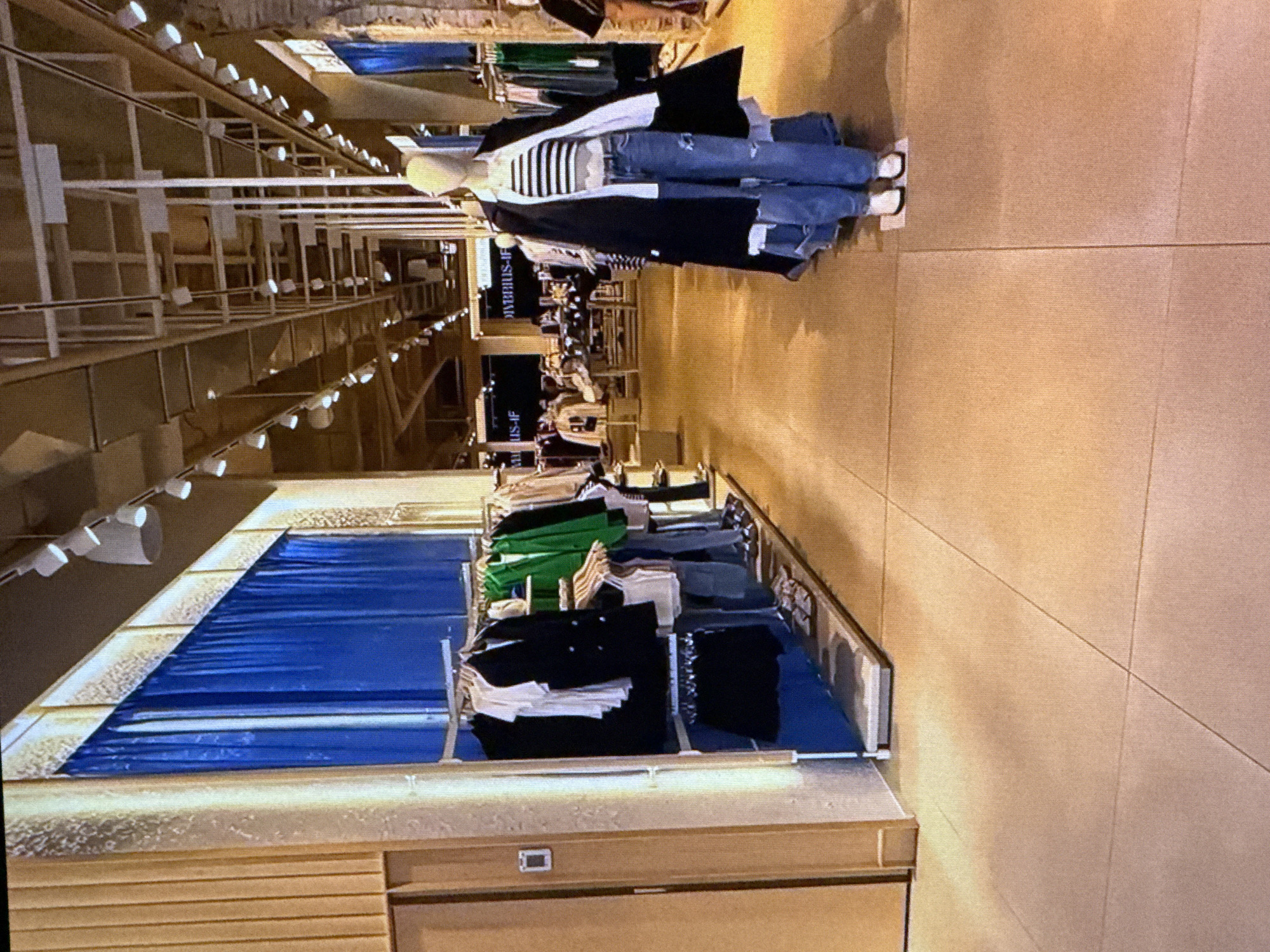 The image depicts the interior of a retail clothing store, likely in a shopping mall, with a focus on displaying merchandise. In the foreground, a mannequin is dressed in a casual outfit consisting of ripped blue jeans, a striped top, and a black coat. The floor is tiled with a light brown color, and the walls appear to be a combination of textured wood paneling and blue fabric curtains. Clothing racks with various garments, including jackets and shirts in colors like black, white, green, and blue, are visible along the walls. The store's ceiling features a grid-like structure with numerous track lights illuminating the space. In the background, more clothing displays and shoppers can be seen, suggesting a busy retail environment. There is no explicit indication of the specific location within Dubai, United Arab Emirates, but the overall aesthetic is consistent with modern retail spaces found in the city.