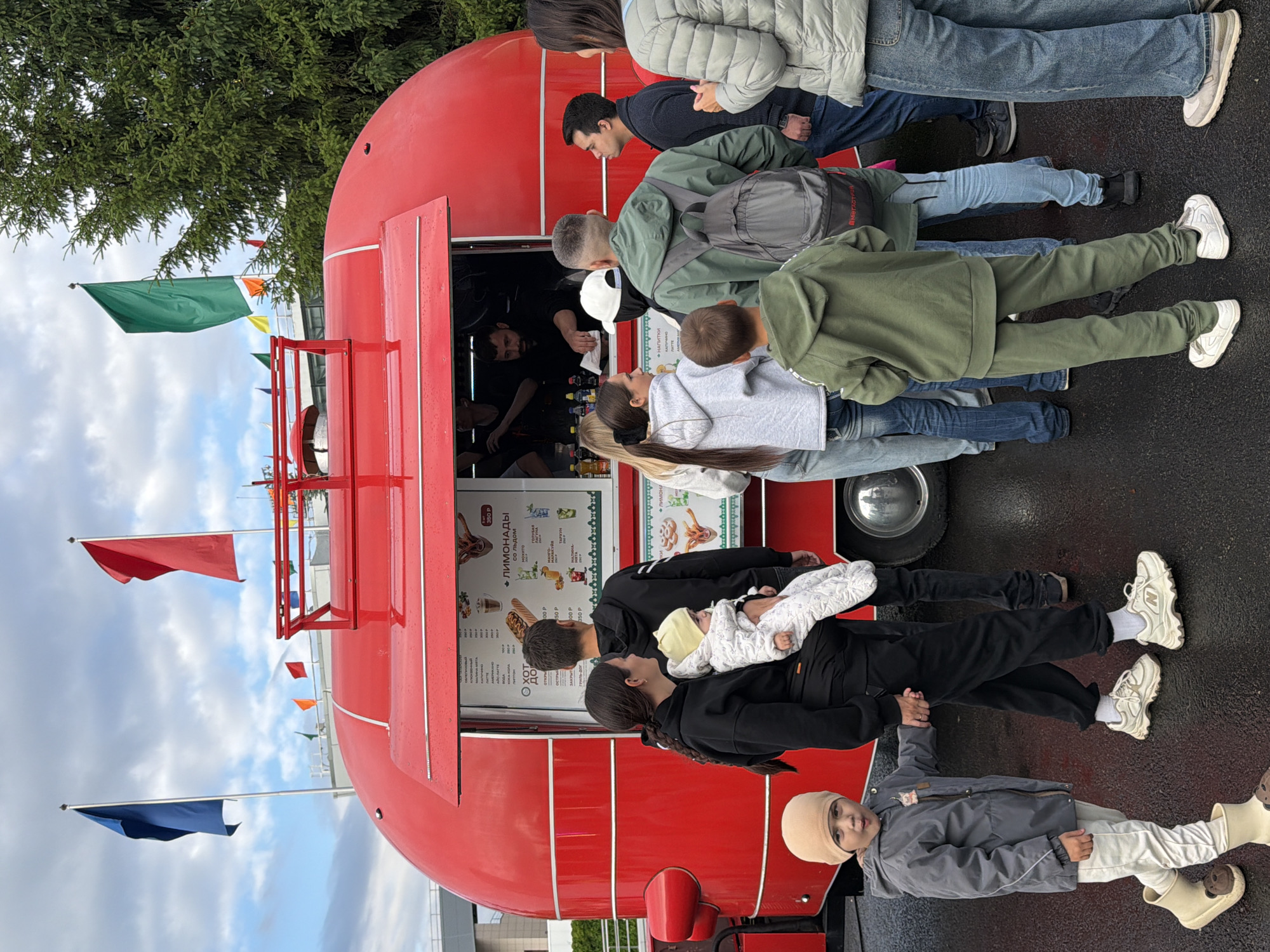 A group of people are gathered around a bright red food truck. The truck has a menu displayed, showing items like "Лимонады" (Lemonades) and various other drinks and food. A man is visible behind the counter, seemingly serving customers. Among the onlookers, there's a woman holding a baby, a young boy in a grey jacket and hat, and several adults in casual attire, some with backpacks. The scene appears to be outdoors, with flags of blue, red, and green flying in the background against a cloudy sky. Trees are also visible in the background. The ground is wet, suggesting recent rain.