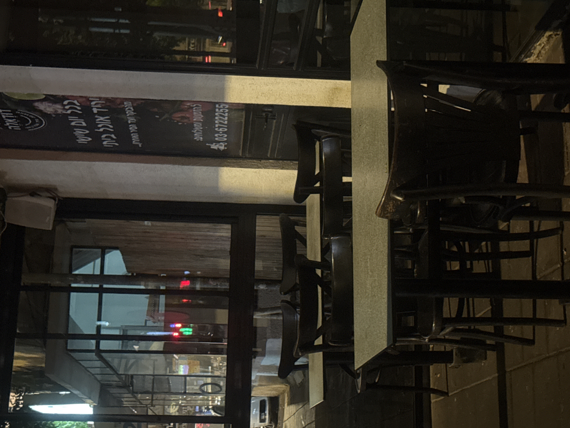 An outdoor restaurant scene in Ramat Gan, Israel features a table and chairs. A sign visible in the background shows a telephone number, 03-6722255, and text in Hebrew. The surrounding environment suggests an urban setting at night.