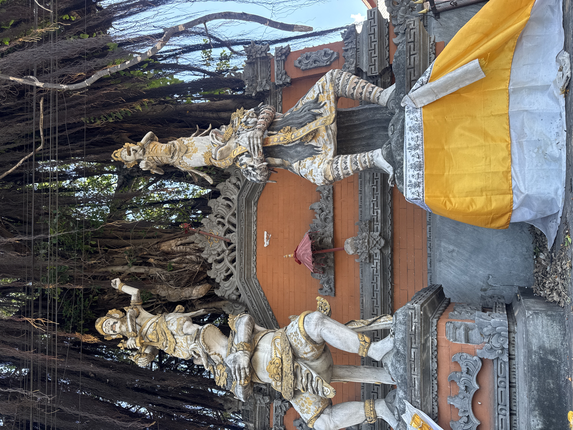 The image shows the entrance to Pura Penataran Agung Batur Sari temple in Kubu, Indonesia. There are two statues, one on either side of the entrance. Both are painted white with gold accents. A yellow and white cloth is draped on the right side. The temple is set in front of a Banyan tree.