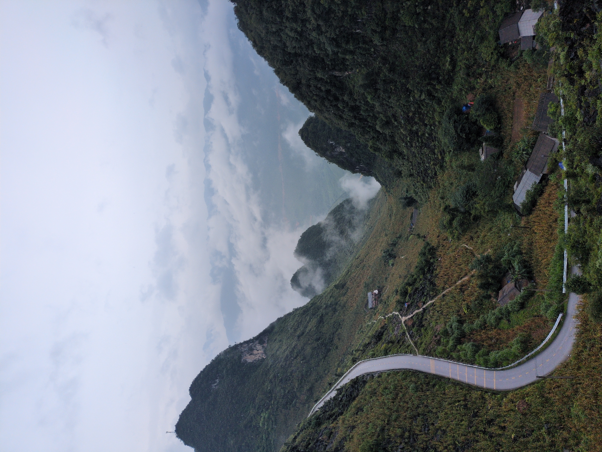 The image captures a scenic view of a winding mountain road in Đồng Văn Commune, Vietnam. The road, paved with asphalt and marked with yellow lines, snakes through a lush, green mountainous landscape. The slopes are covered in dense vegetation, with patches of dry grass and scattered trees. Small clusters of houses with dark roofs are visible nestled on the hillsides, indicating a rural settlement.
In the background, a dramatic vista unfolds with several peaks piercing through a layer of thick, white clouds. The sky is overcast with a pale, grayish hue. The overall atmosphere is serene and somewhat mystical, emphasizing the raw beauty of the natural terrain. The perspective suggests the photograph was taken from a higher elevation, looking down onto the road and the valley. There are no people or animals visible in the image.