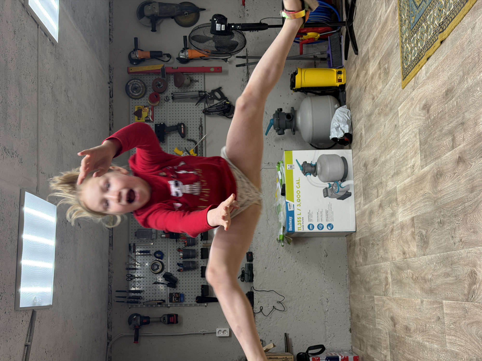A child in a red shirt is holding their leg in the air in a room, possibly a garage or workshop, with tools mounted on the wall behind. The room has a gray concrete wall and a light fixture is mounted in the corner of the wall. A box for a water filter and a fan are visible. The photo does not contain the information about the location, so the location cannot be confirmed.