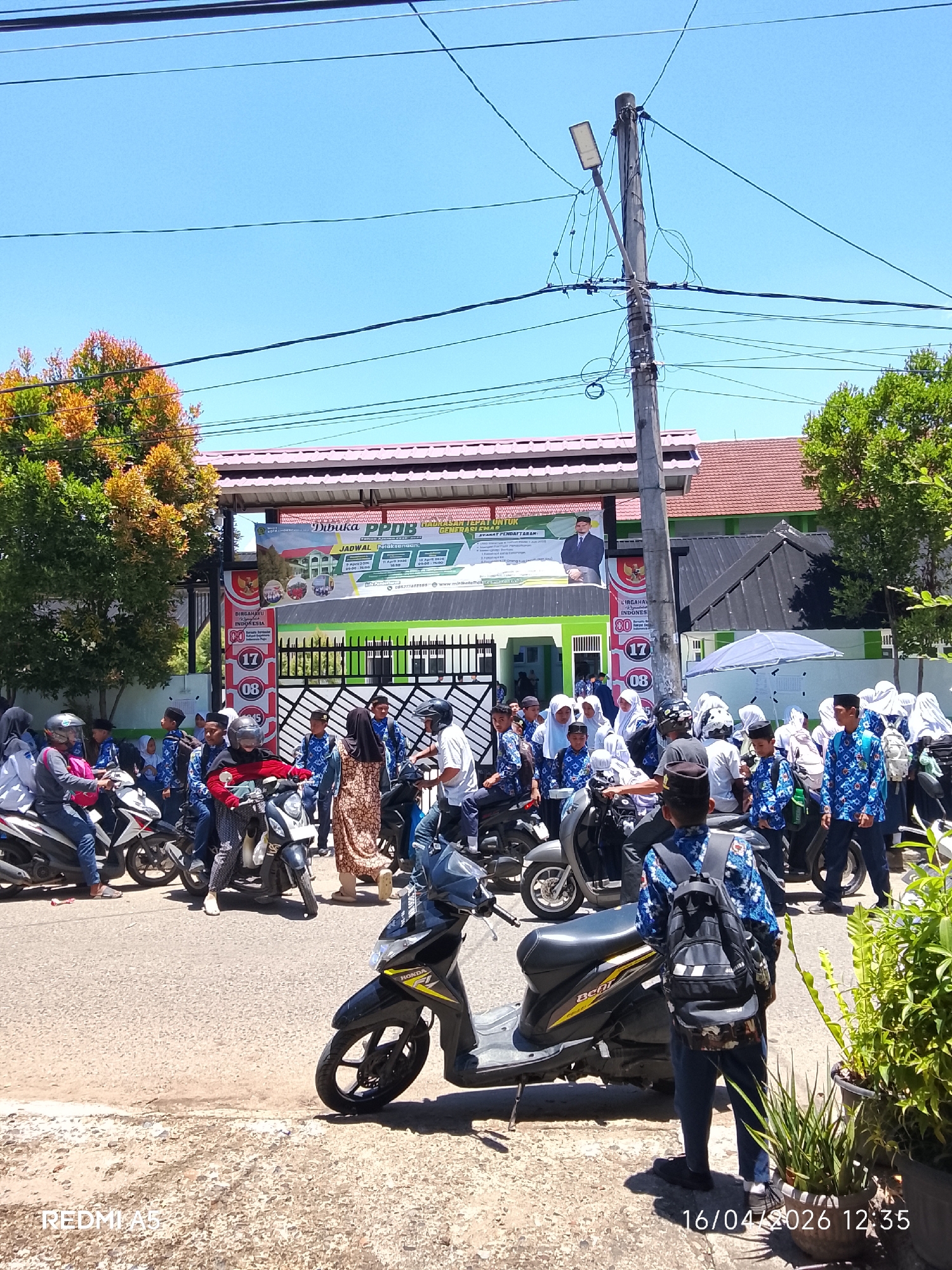 The image depicts a scene in Lhokseumawe, Indonesia, in front of a building with a banner announcing the opening of PPDB (Peserta Didik Baru) which refers to the new student admissions. The banner also contains the official symbol for the 80th Indonesian Independence Day, indicating an upcoming celebration. A group of people, mostly students dressed in similar blue patterned uniforms, is gathered on the street. Many are on motorcycles, while others are standing or walking. The environment suggests a school or public institution with a gate, plants, and overhead electrical wires and poles. The date stamp shows "16/04/2026 12:35."