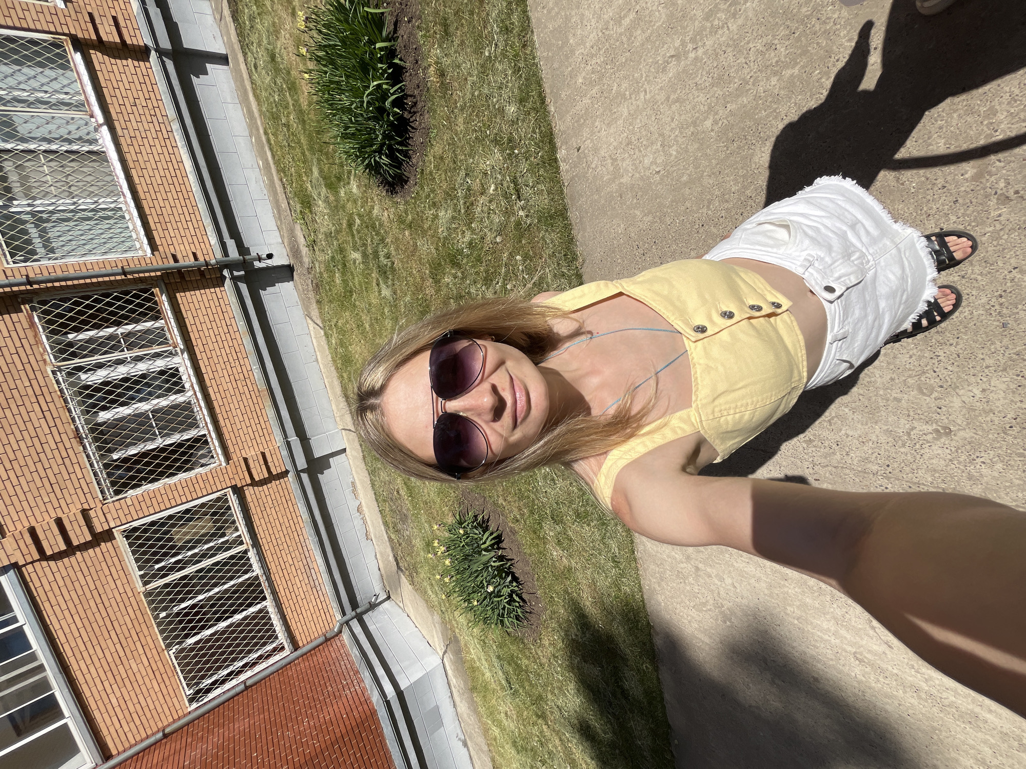 A high-angle shot shows a woman in front of a brick building with barred windows, potentially in the city of Chaikovsky, Russia. The woman wears sunglasses, a yellow top, and a white denim skirt, with black sandals on her feet. She appears to be taking a selfie, her arm extended toward the camera. A strip of grass and small plantings separate her from the building. A shadow, likely her own, is cast on the concrete ground. The scene suggests an outdoor urban or semi-urban setting on a sunny day.