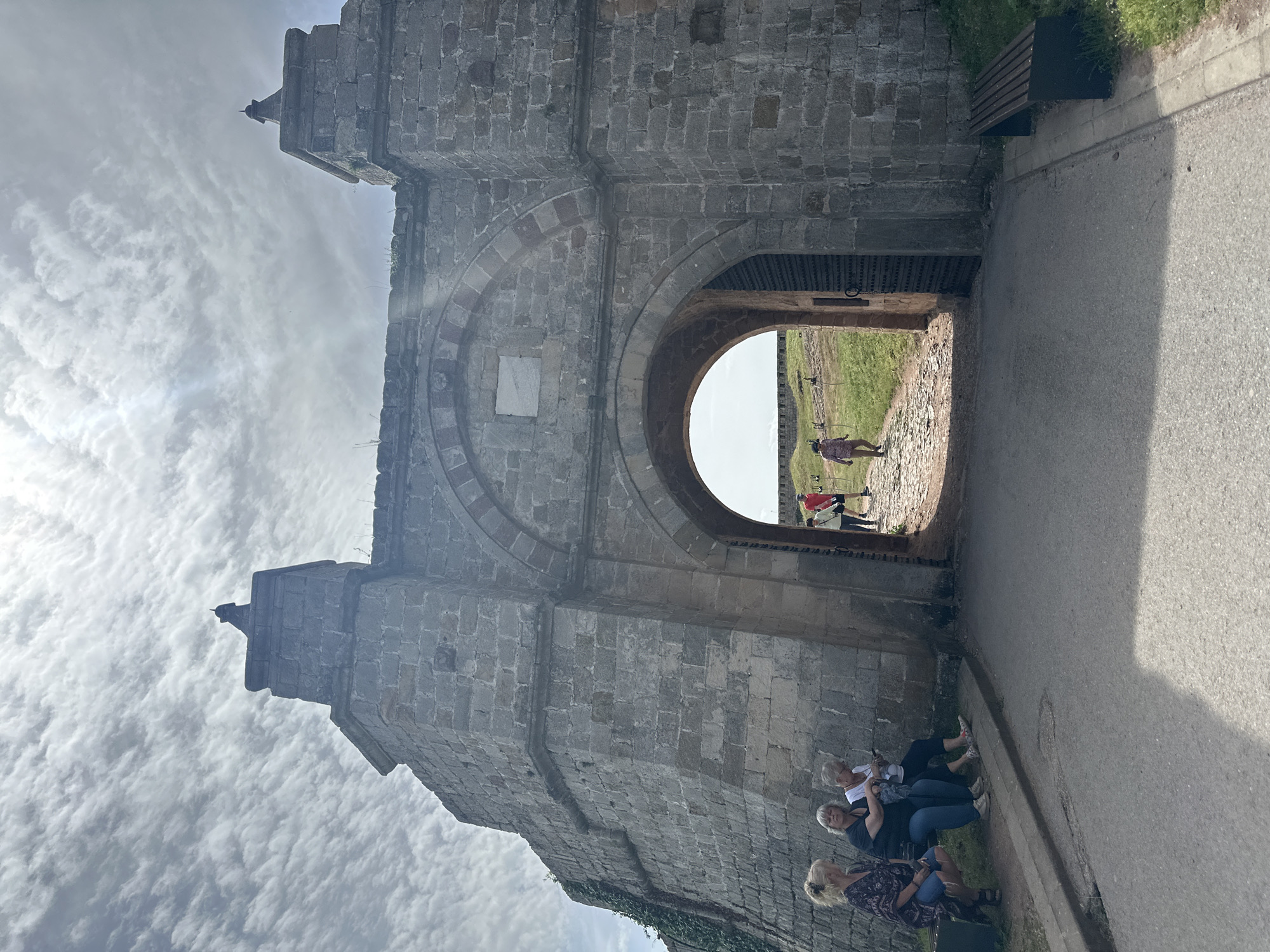 The image shows the main gate of Belogradchik Fortress in Bulgaria, constructed of light gray stone. The gate is a large archway with a smaller circular arch inside. A group of people is sitting on the ground to the right of the gate, and several people are visible in the background, walking through the gate. The sky is visible above the fortress, with light, wispy clouds. A stone path leads to the gate, and there is grass and a bench visible near the path.
