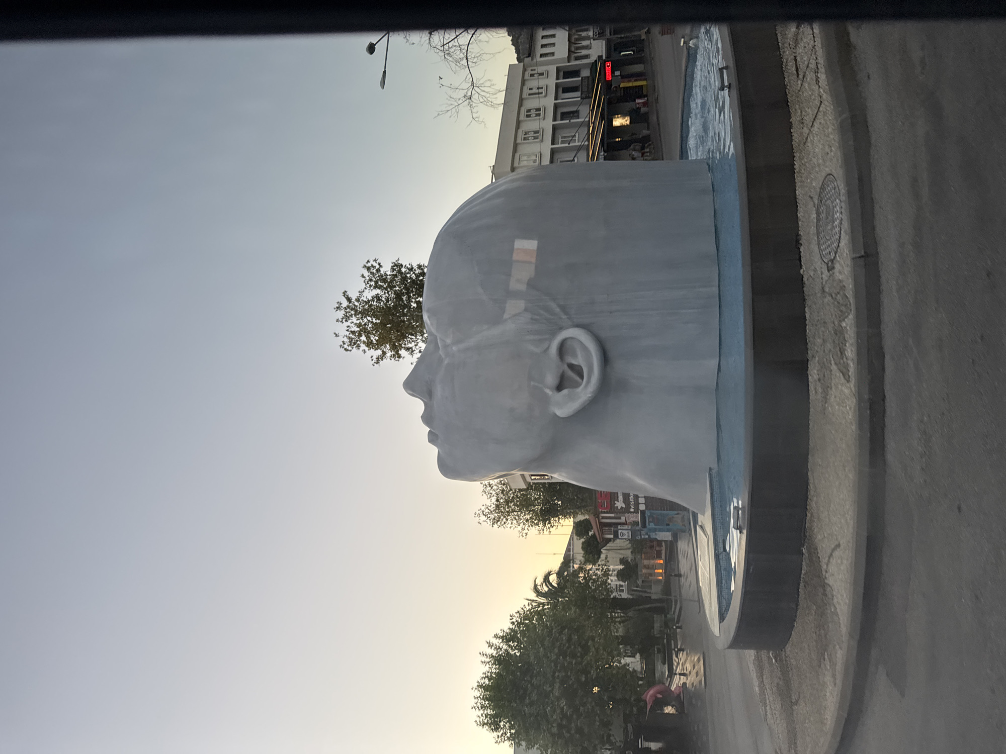 The image captures an outdoor scene in Kemer, Turkey, featuring a large, grey, abstract sculpture of a head tilted back and facing upwards. The sculpture appears to be part of a fountain, with water flowing from its base into a circular pool. The sculpture is situated in what seems to be a public square or intersection, with a paved ground surrounding the fountain.
In the background, buildings with multiple stories and windows are visible. There are trees and streetlights present in the scene. The sky is a gradient of light blue and pale yellow, suggesting it might be dawn or dusk. A dark, vertical element, possibly a pillar or a portion of a vehicle, partially obstructs the right side of the frame.
Further elements in the background include what appears to be a street with some pedestrian activity. A pink object, possibly a statue or signage, is seen to the left of the fountain. Reflections of windows are visible on the surface of the sculpture.