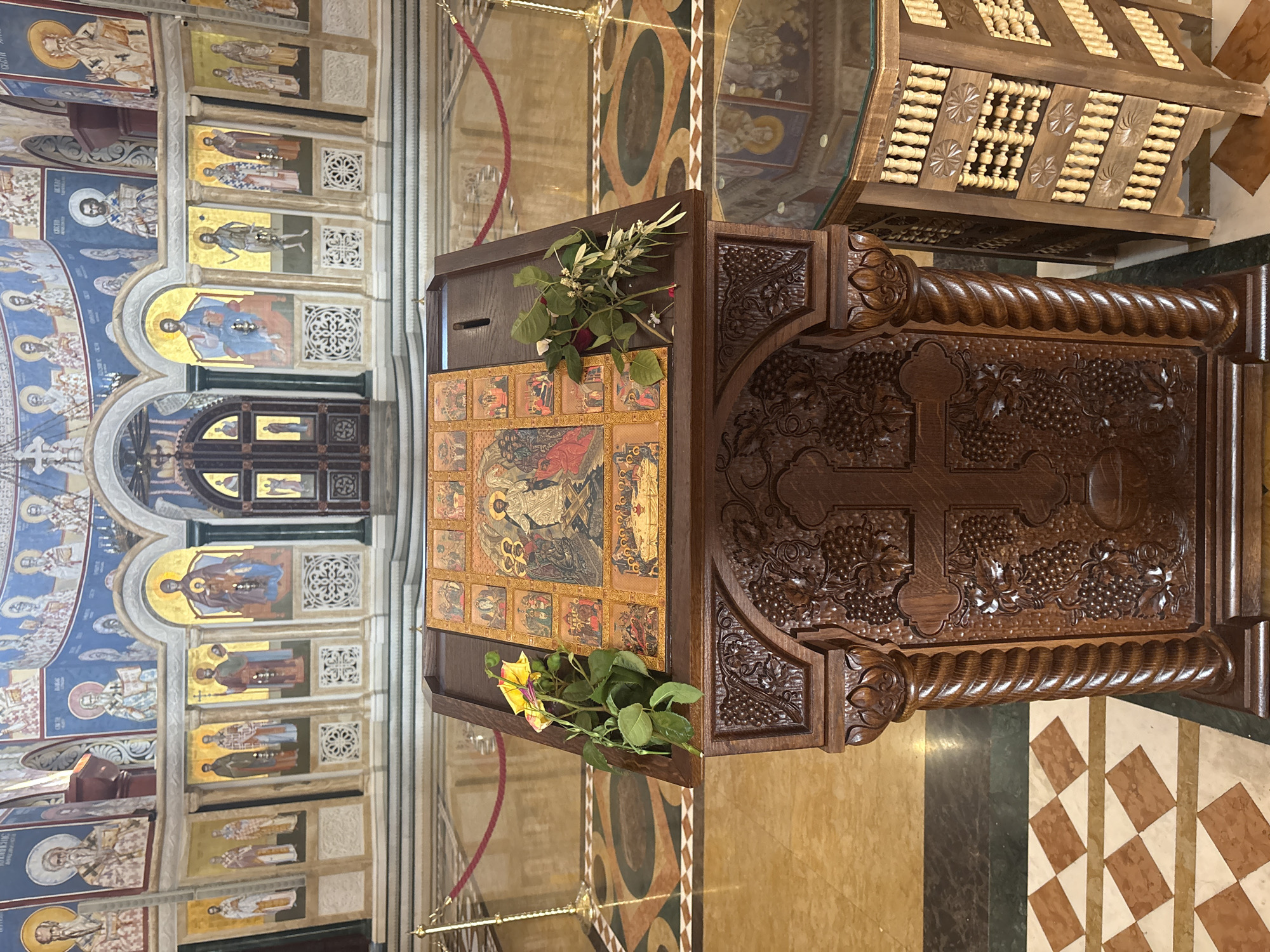 The image depicts the interior of a church, likely in Bar, Montenegro, based on its architectural style and religious iconography. The scene focuses on a large, ornately carved wooden lectern or pulpit, prominently featuring a cross and grape vine motifs, set on a marble floor. The lectern is adorned with a series of framed religious icons and has arrangements of flowers placed on it. Behind the lectern, the church walls are covered in frescoes depicting religious figures. The ceiling is arched and also features elaborate religious paintings. In the background, there is a closed wooden door. The overall environment suggests a formal and sacred space.