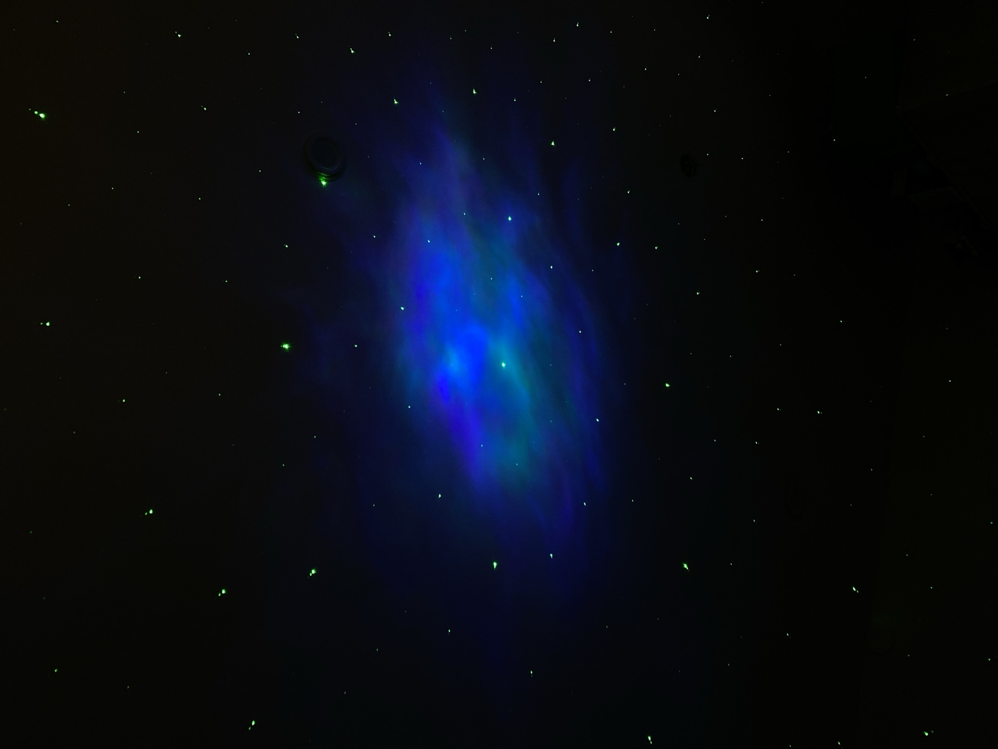 The image depicts a dark, indoor space resembling a simulated night sky. A nebulous, blue-green shape dominates the center, suggesting a projected or illuminated effect. Small, green points of light are scattered across the black background, mimicking stars. Near the top left, a circular object appears, possibly a speaker or a light fixture. The exact location within Votkinsk, Russia, cannot be determined from the image alone. No people are visible, and the primary objects are the simulated nebula, stars, and a fixture on the ceiling. The interaction is between light and the environment to create the effect of a night sky.