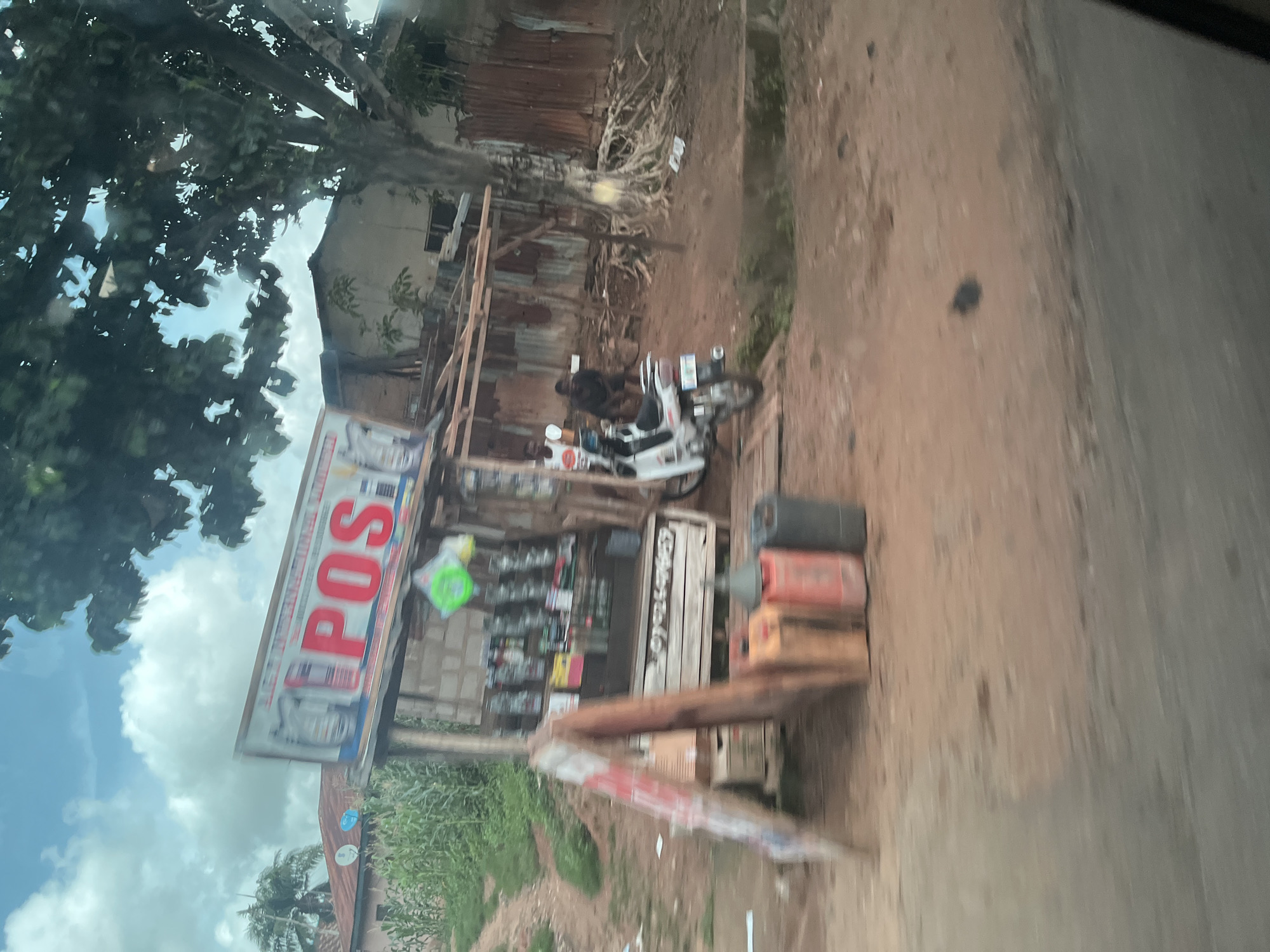 The image shows a roadside stall in Akwanga, Nigeria. The stall has a sign that says "POS" in large red letters. It is stocked with various items, including bottled drinks and other products. In front of the stall, there is a white motorbike and several large jerrycans filled with what appears to be fuel. Two men are visible near the stall; one is standing next to the motorbike, and the other is sitting. The scene is set against a backdrop of houses, vegetation, and a large tree. The ground is a reddish-brown dirt. The sky is visible, with clouds and some blue sky. The image appears to have been taken from a moving vehicle.