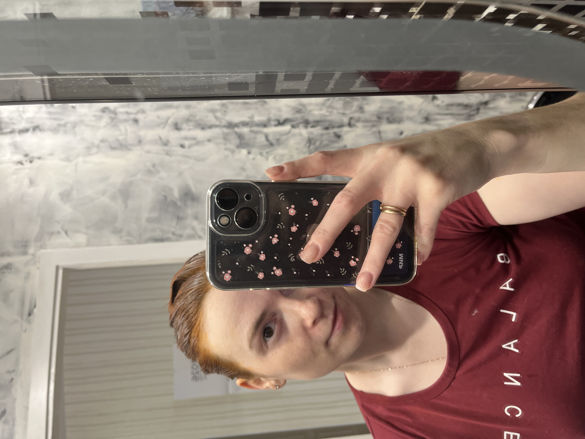 A person with fair skin and red hair is taking a selfie in front of a mirror. The phone is in the foreground, with a black case adorned with small pink floral designs. The person is wearing a dark red t-shirt with the word "BALANCE" printed vertically in white. The mirror reflects a bathroom setting, with walls covered in a light gray patterned tile and a white doorframe. The location is potentially a bathroom within the city of Chaikovsky, Russia, as specified by the user.
