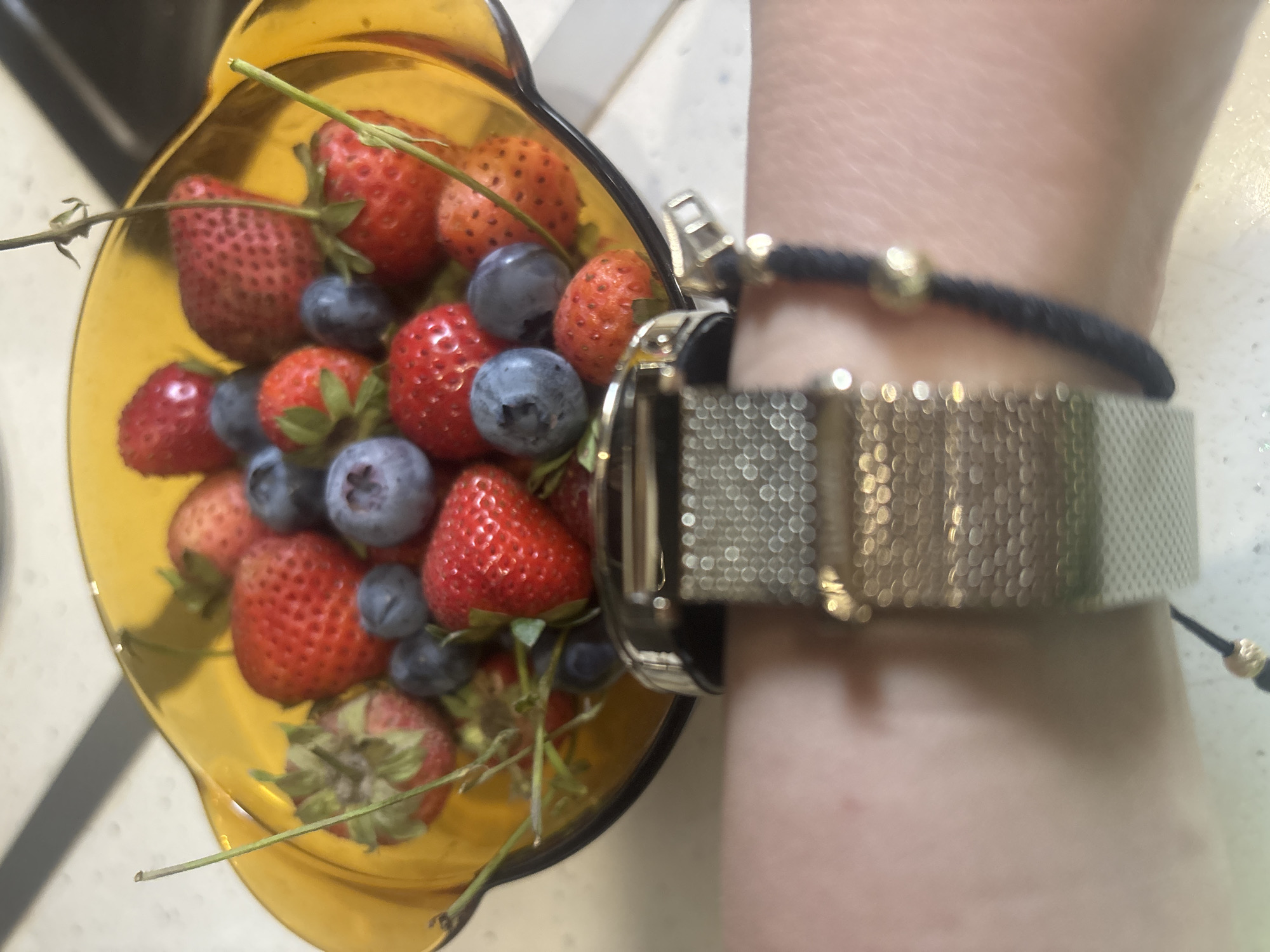 A close-up, eye-level shot shows a bowl of fresh strawberries and blueberries. The bowl is amber-colored glass and is filled to the brim with the red and blue berries. Several green stems from the strawberries are visible, adding a touch of freshness to the arrangement. In the foreground, a person's wrist is visible, adorned with a silver mesh watch and a thin black bracelet with a gold bead. The watch is clasped and appears to be made of stainless steel. The background is blurred, but appears to be a countertop with a speckled texture. No specific location in Burgas, Bulgaria is identifiable from the image.
