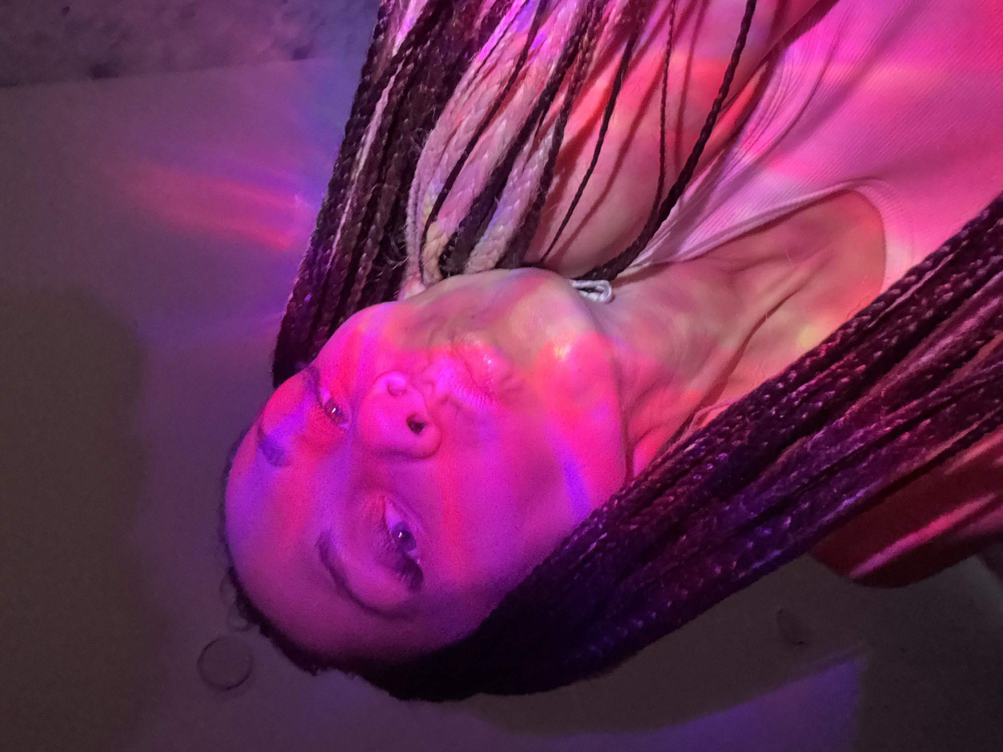 An image captures a person, likely female, from the chest up, with their head tilted backward and looking towards the camera. The dominant features are the person's face, neck, and upper chest, framed by numerous dark, possibly braided, hair strands. Some of the braids show lighter, possibly colored, sections.
The scene is dramatically lit by vibrant artificial lights, casting strong pink and purple hues across the person's skin, hair, and light-colored top. These lights create colorful reflections and shadows, particularly visible on the face and décolletage. A subtle, light-colored detail, possibly a necklace or part of the clothing, is visible near the collarbone.
The background is largely dark and indistinct, with soft gradations of purple and pink light, suggesting an indoor setting with atmospheric or mood lighting, possibly in an entertainment venue or a private space designed for relaxation. The person appears to be in a relaxed posture, engaging directly with the camera. Their expression is neutral, with eyes open, indicating they are posing for the photograph. There is no visible text in the image. The image is set in Bor, Russia.