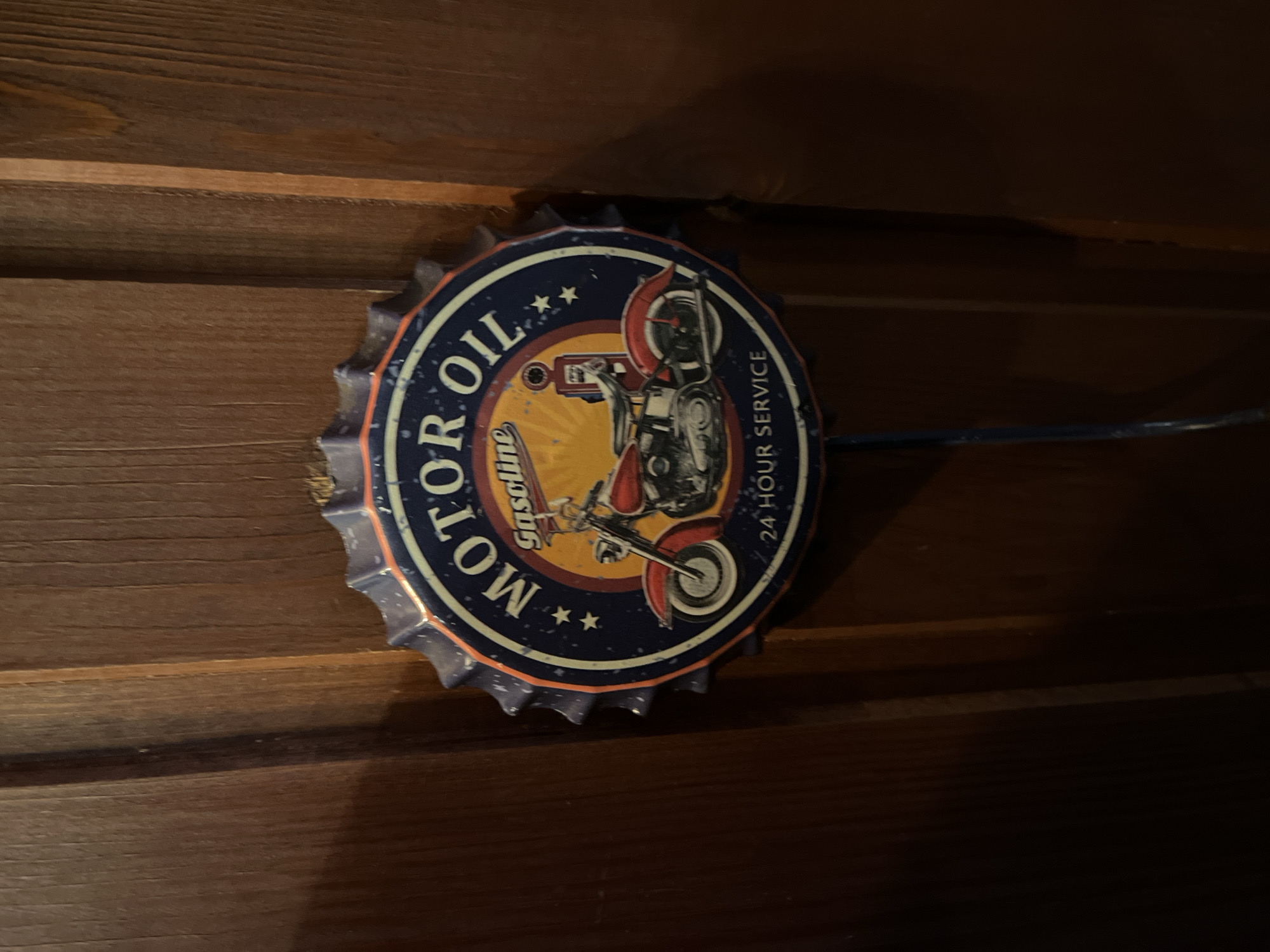 The image shows a vintage-style "Motor Oil Gasoline 24 Hour Service" metal sign affixed to a dark, horizontally-paneled wooden surface. The sign is circular with a bottle cap-like edge. The background is dark and dimly lit, suggesting an indoor setting.
There is no indication of the location being Petropavlovsk-Kamchatsky, Russia, or of any people, actions, or interactions.