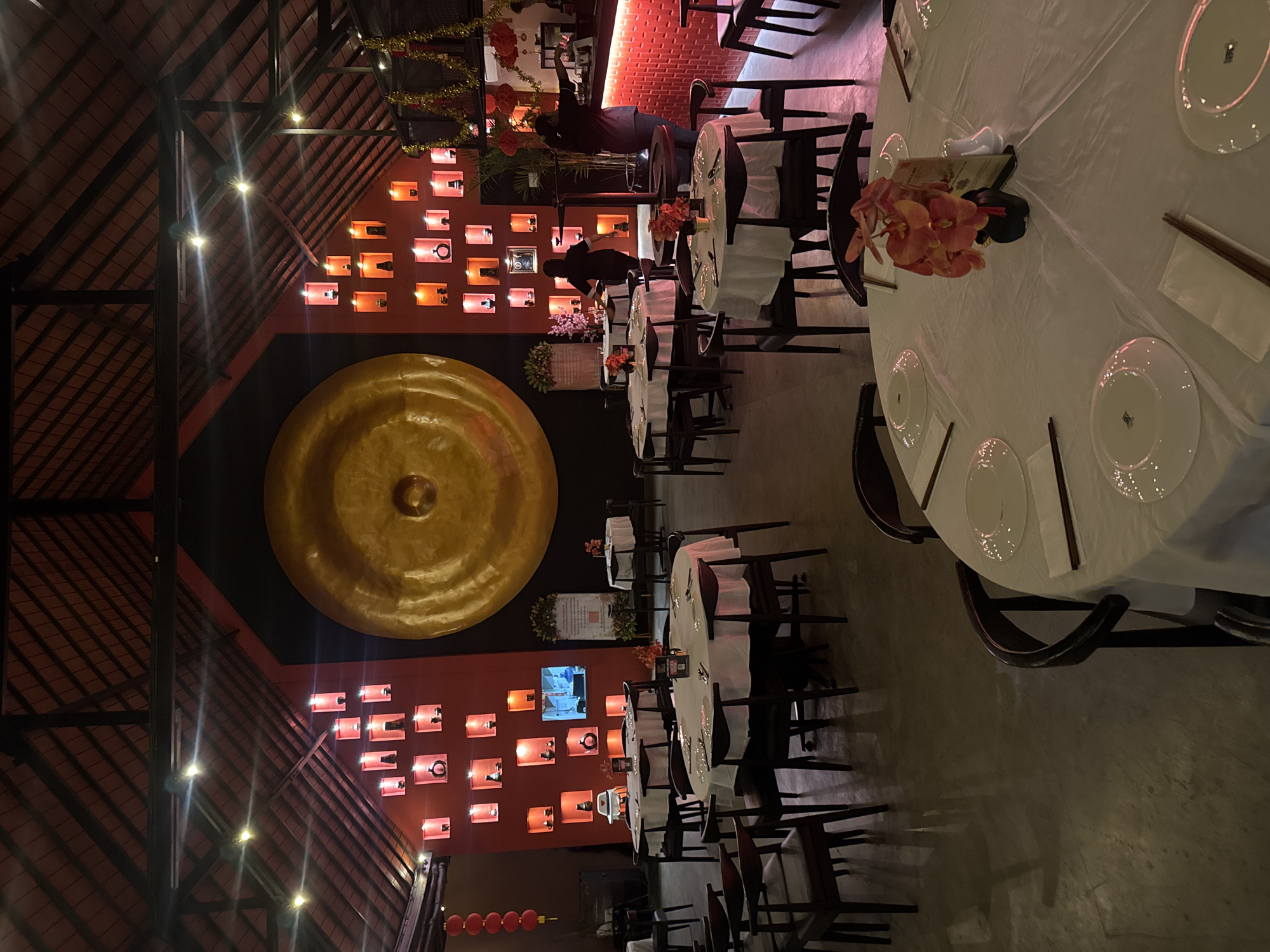 The image depicts the interior of a restaurant, likely an Asian establishment, given the decor and the presence of a large gong. The scene is set at night, with warm lighting from lamps and accent lights creating a cozy ambiance. Several round tables are set with white tablecloths, plates, and chopsticks, indicating that patrons are either dining or about to dine.
Two individuals are visible, appearing to be staff members. One, dressed in dark clothing, is standing near the bar area, seemingly attending to something. Another person, also in dark attire, is bent over a table, possibly arranging place settings or serving food. In the background, shelves are adorned with various decorative items and bottles, illuminated by red lighting.
Notable details include the rustic wooden ceiling structure, the prominent golden gong on the wall, and rows of red lanterns hanging outside. The overall atmosphere suggests a lively and inviting dining experience. While no specific text is clearly legible, menu cards are present on some tables. The setting is in Kuta, Indonesia.