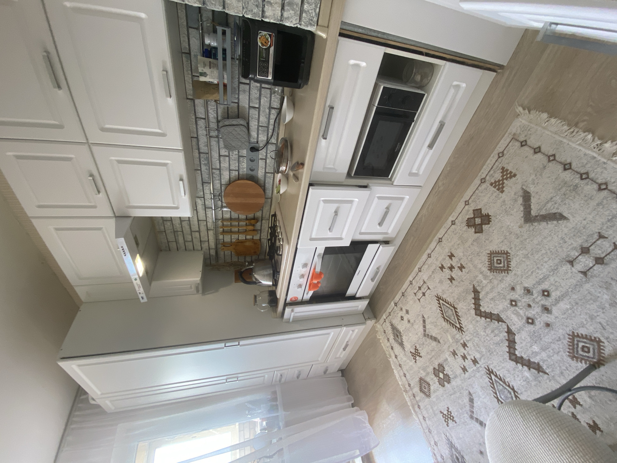 This is a medium-shot, eye-level, full-frame photograph of a small, modern kitchen. The kitchen is outfitted with white cabinetry, a light-colored countertop, and a brick-patterned backsplash. Appliances visible include a gas stovetop, an oven with orange oven mitts inside, a microwave, and an air fryer. A patterned rug covers a portion of the light-colored wood floor. A window with sheer white curtains is visible on the left side of the frame. A tall white cabinet unit is located to the left of the stove. The lighting in the image appears to be natural light from the window, supplemented by overhead lighting. The scene depicts a clean and organized kitchen space.