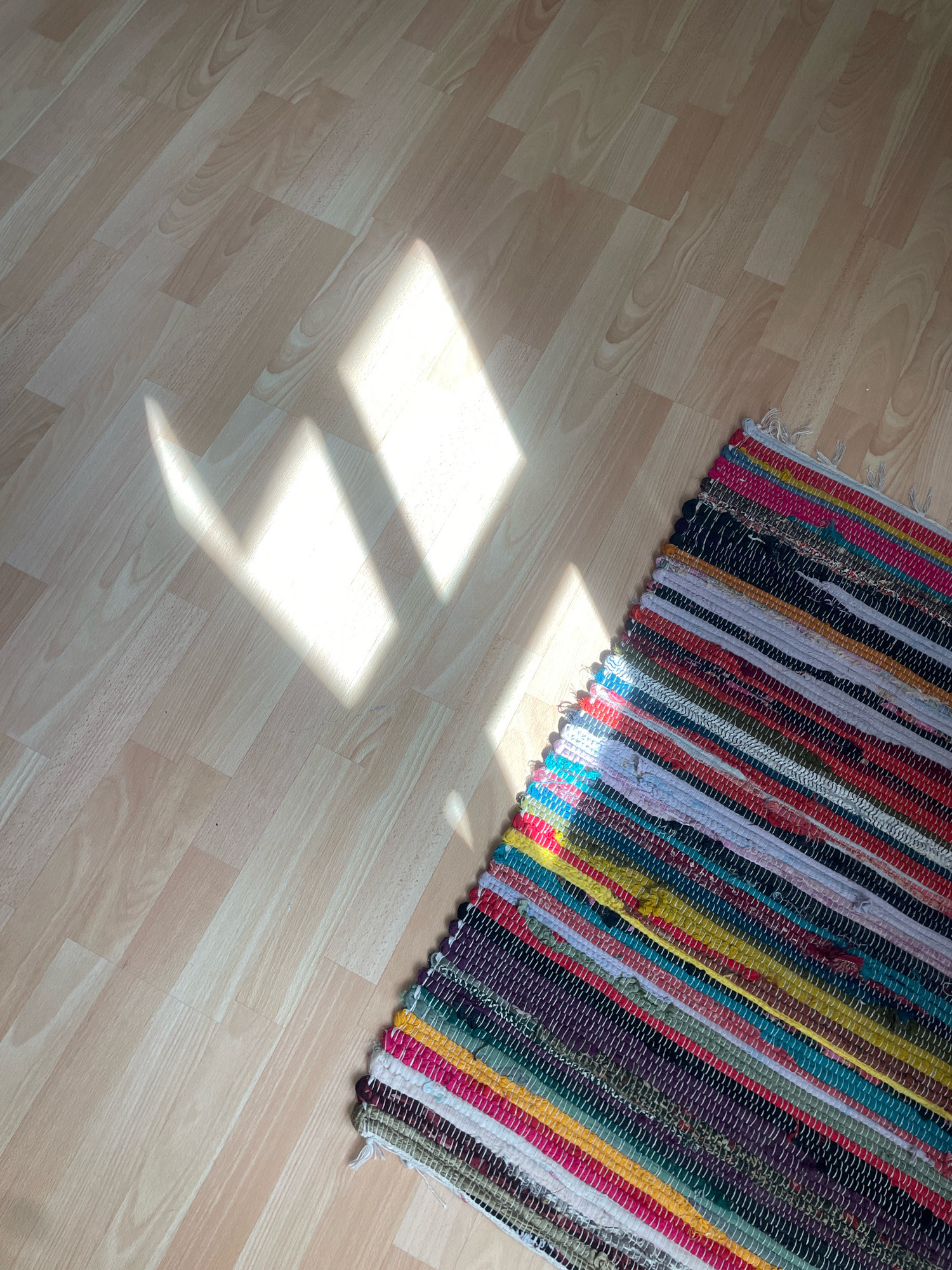 The image shows a colorful, striped rag rug lying on a light-colored wooden floor. Sunlight is casting geometric shadows on the floor. The scene appears to be an indoor space. No people are visible in the image. The specific location in Atarfe, Spain cannot be determined from the visual information provided.