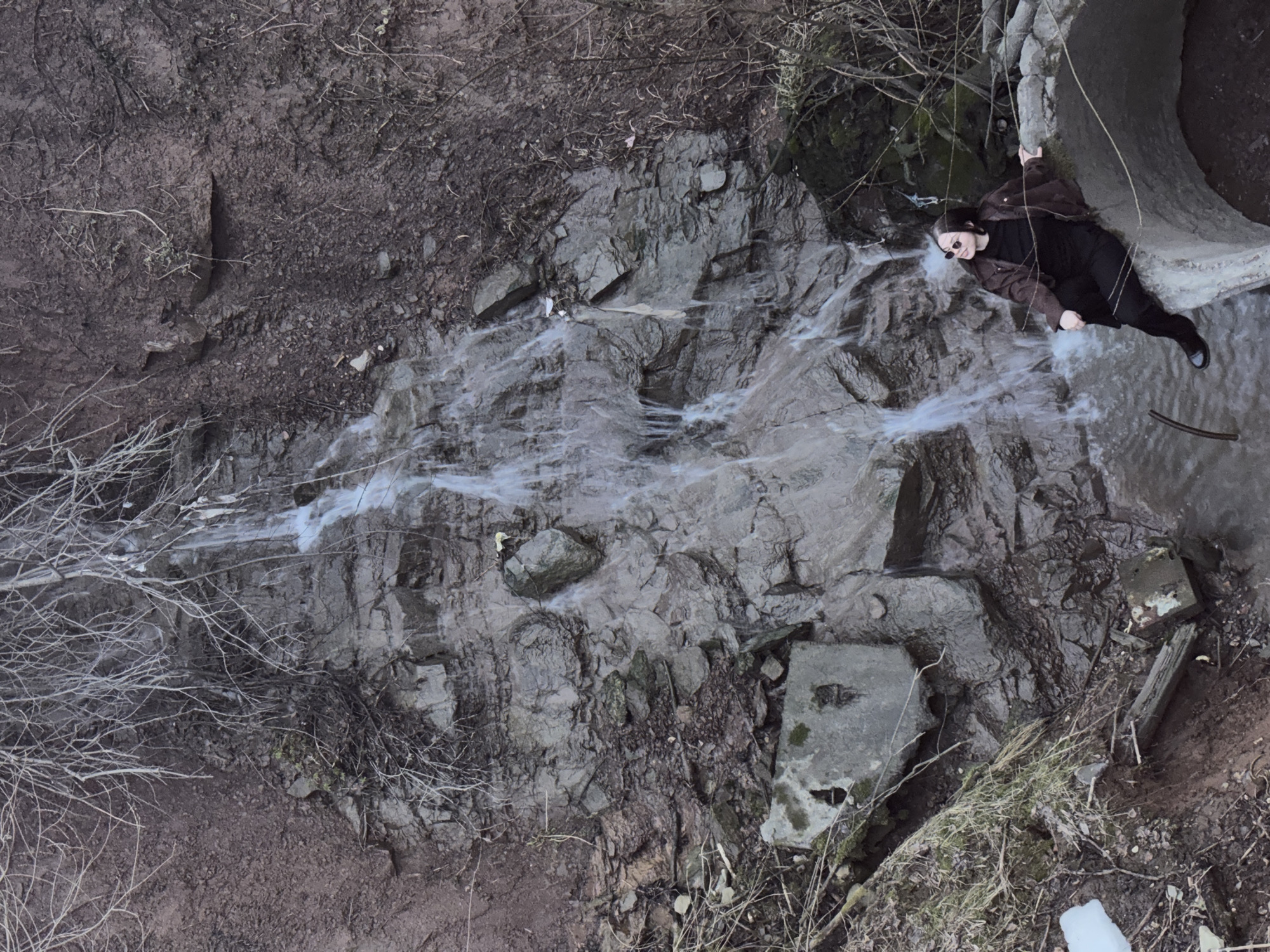 A high-angle shot depicts a landscape in Perm, Russia, dominated by a rocky stream bed with flowing water. A woman in dark clothing and sunglasses is positioned on the right side of the frame, clinging to a concrete structure or pipe outlet. She appears to be leaning outwards, with one leg dangling over the edge. The surrounding environment is characterized by dirt, rocks, dry branches, and a concrete block partially covered in moss. A small white plastic object is visible on the lower right. The color palette consists of muted earth tones.