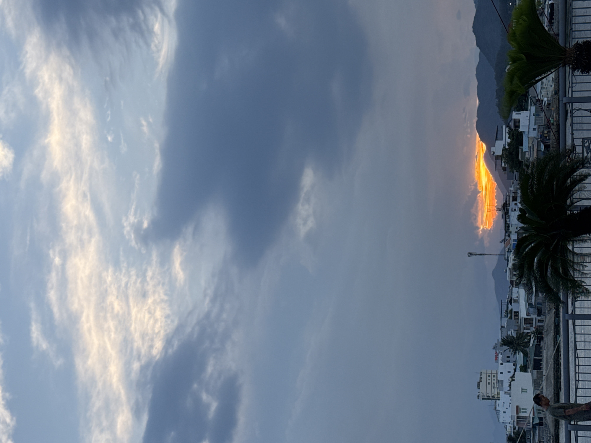 The image shows a sunset over a city skyline in Nha Trang Ward, Vietnam. The sky is filled with dramatic clouds, ranging from deep grey to bright orange where the sun is setting behind a mountain range. In the foreground, there is a body of water with a railing along its edge. A man in a green shirt walks along the railing, looking down. Beyond the railing, there are buildings with white facades, some appearing to be apartments or hotels, interspersed with palm trees. The overall scene depicts a coastal city at dusk.