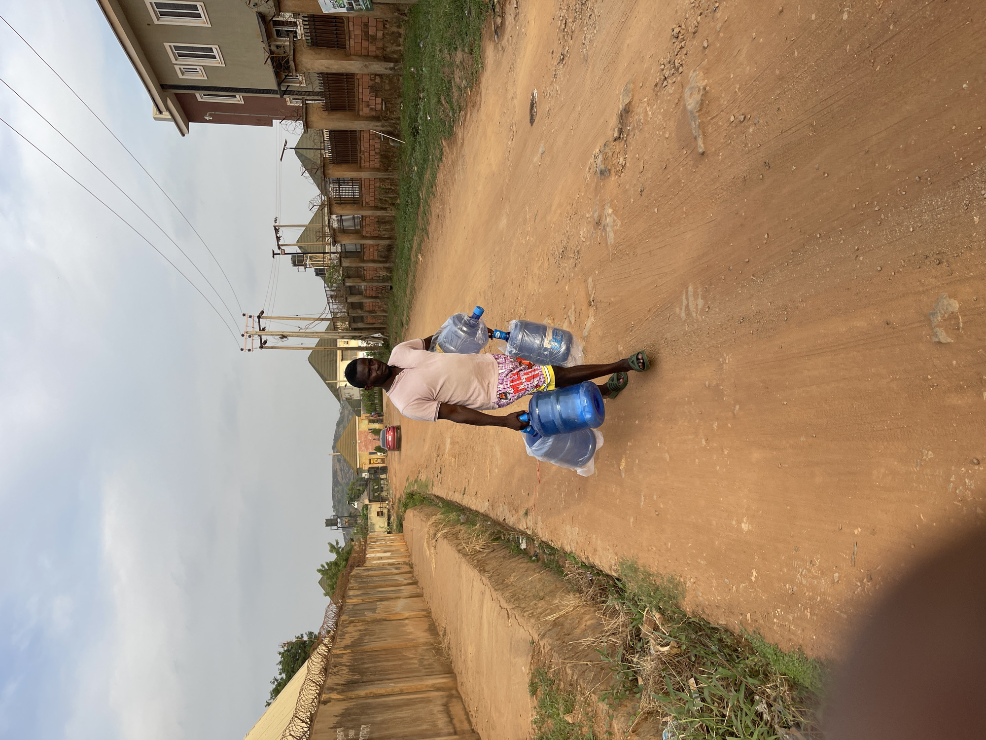The image captures a daytime scene in what appears to be a residential area. The primary subject is a man walking on an unpaved, reddish-brown dirt road. He is carrying three large blue water dispenser bottles; two are held in his left hand and one in his right, suggesting they are heavy. He wears a light-colored t-shirt, multi-colored patterned shorts, and green sandals.
The road is dusty with scattered small stones and faint tire tracks. Along the left side of the road runs a concrete barrier or wall, topped with barbed wire fencing. Beyond this barrier, there's some green vegetation. On both sides of the road, various multi-story buildings are visible, constructed with concrete and bricks, some featuring white-framed windows.
In the background, power lines stretch across the sky, connected to wooden utility poles. The sky is overcast, indicating a cloudy day. Distant hills or mountains can be seen on the horizon. The overall setting appears to be a developing urban or semi-urban environment, characteristic of places like Abuja, Nigeria, where the transport of water in large containers is a common activity. Visible text on a distant sign and on the concrete barrier is too faint and distant to be legible.