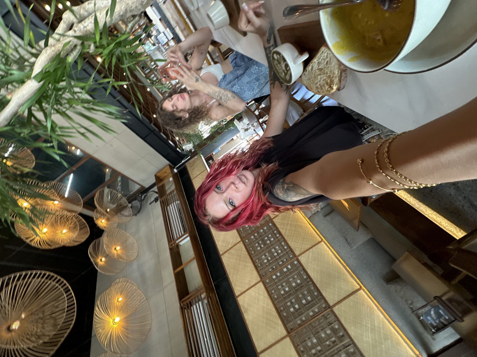Two women are pictured sitting at a table in a restaurant in North Kuta, Indonesia. The first woman, with curly brown hair, is using a smartphone. The second woman, with pink hair and gold bracelets, is taking a selfie. A bowl of soup, a cup, and a slice of bread are on the table. Behind them is a wall with decorative light fixtures, and there is a plant in the background.