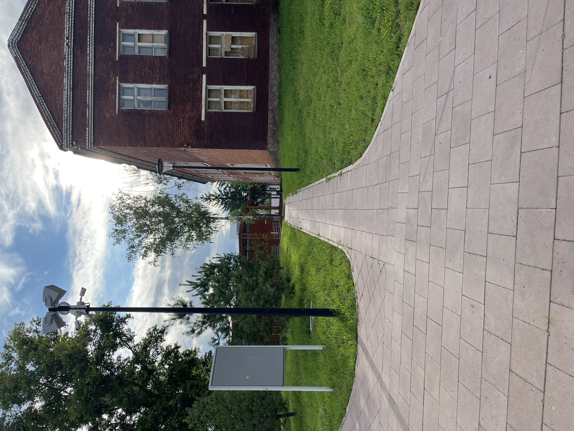 The image shows a paved walkway leading towards a red brick building with boarded-up windows. The walkway is made of rectangular concrete pavers and curves to the right. To the left of the walkway, there is a patch of green grass with a gray notice board on a stand and a tall lamppost with multiple lights and cameras. Further to the left and in the background, there are trees. The building appears to be old and somewhat neglected. The sky above is partly cloudy with sunlight filtering through. The scene is likely an outdoor area in a city, possibly a park or a public space adjacent to the building. It is not possible to determine the specific location in Blagoveshchensk, Russia from the image alone.
