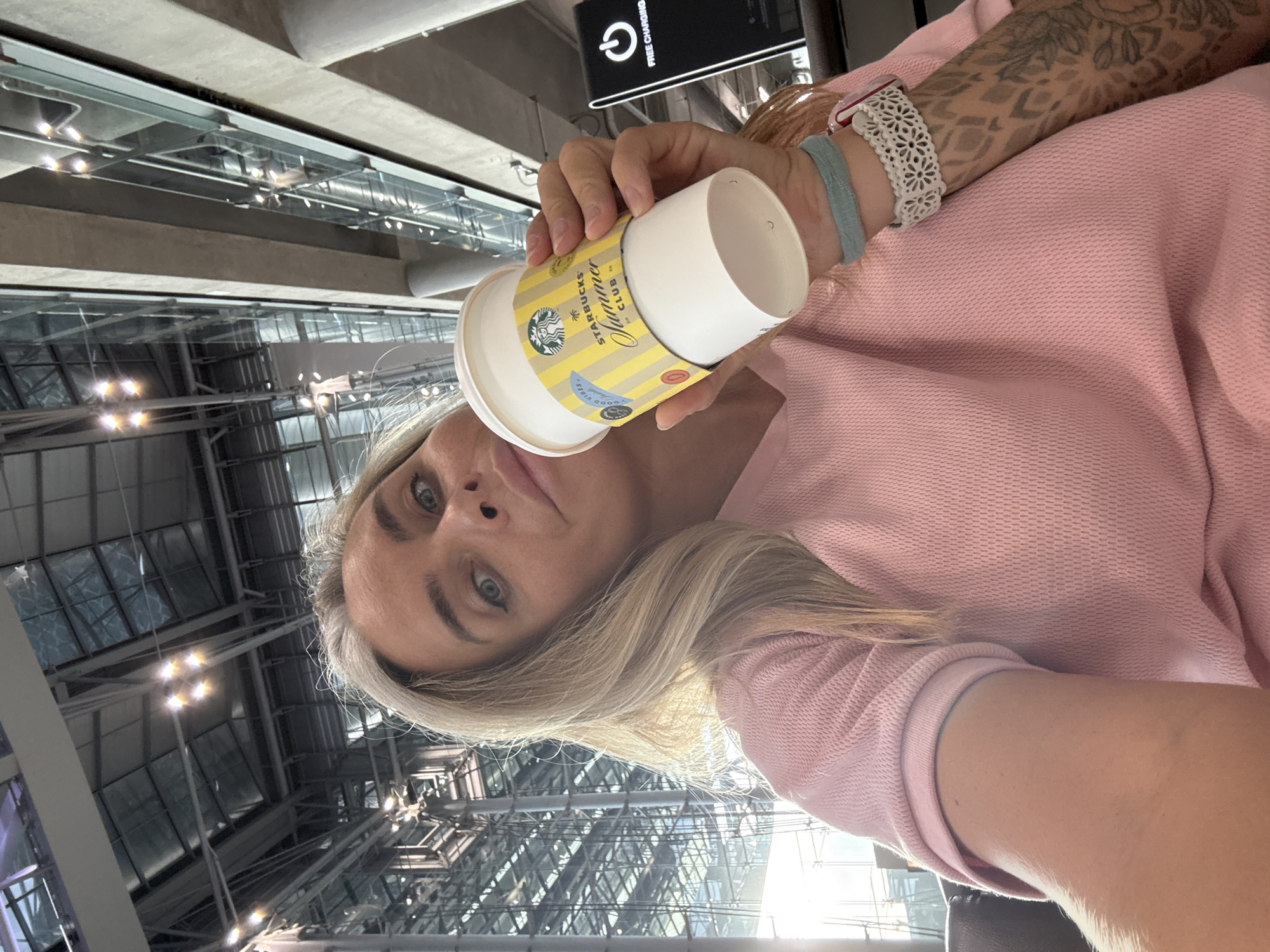 A woman with blonde hair and light green eyes is holding a Starbucks cup in her right hand. She is wearing a pink top. She has tattoos on her right arm and is wearing bracelets. The background appears to be an indoor public space with a high ceiling, exposed metal beams, and large windows. A sign with a charging symbol and the words "FREE CHARGING" is visible to the right. This location is the Suvarnabhumi Airport, located in Nong Prue Subdistrict, Bang Phli District, Samut Prakan Province, Thailand.